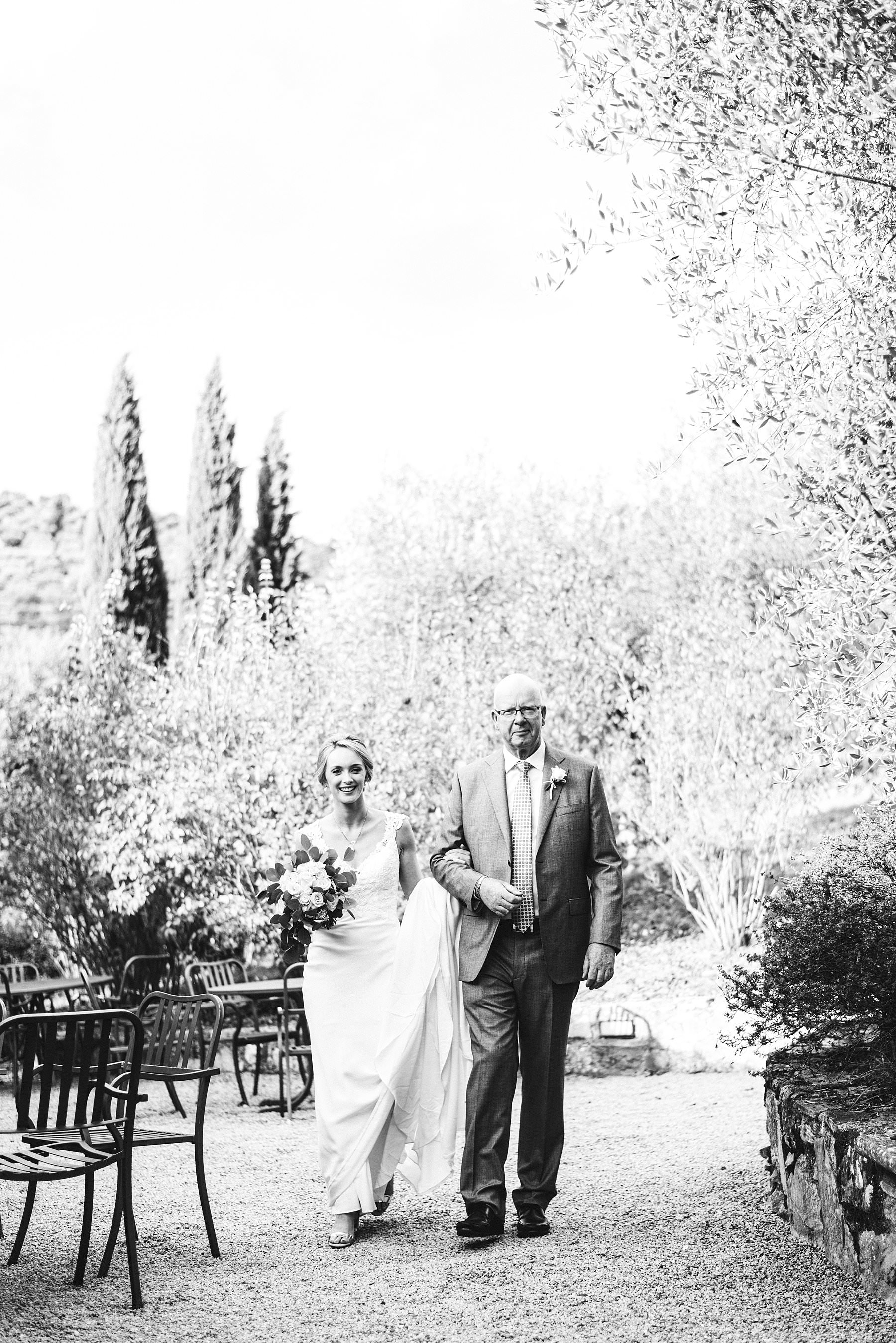 A special international wedding in the countryside of Umbria, Italy. Elegant Australian bride Jennifer walks with father in the symbolic ceremony held in a courtyard close to the Villa Monte Solare, surrounded by greenery and decorated with delicate flowers