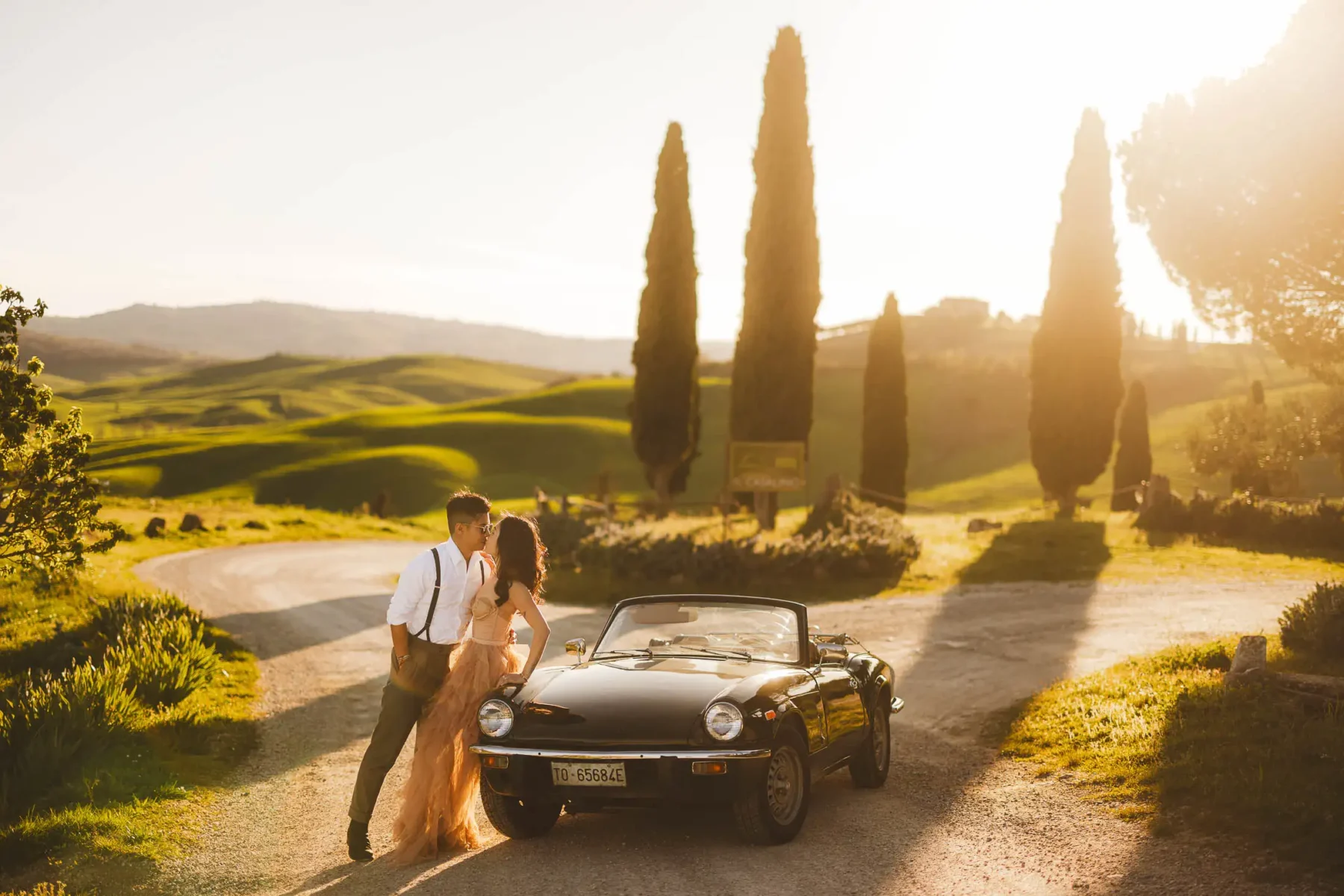 Lovely and elegant pre-wedding engagement couple photo shoot in the countryside of Pienza in the Val d'Orcia with a old Vintage Car during golden hour and evocative cypresses and rolling hills scenery