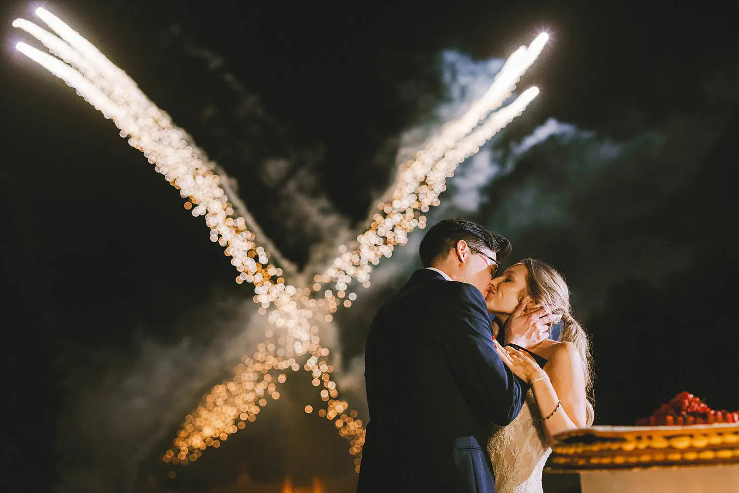 Bride and groom kiss with fireworks illuminating the night sky at luxury wedding at Castello di Celsa in Tuscany