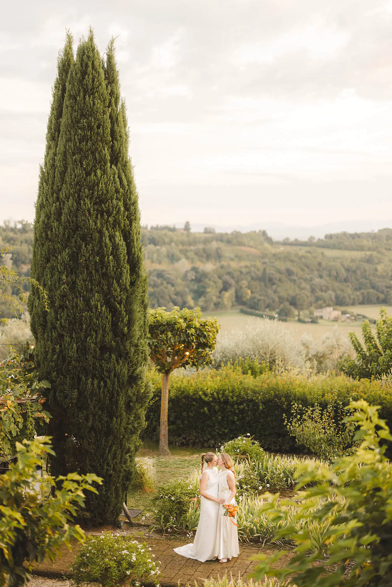 Two brides sharing a quiet kiss surrounded by the rolling Tuscan countryside during their same sex destination wedding at Borgo di Petrognano, where nature, intimacy and timeless beauty come together
