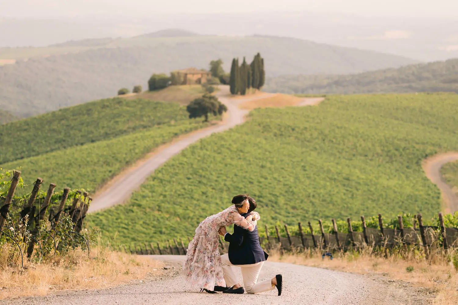 A couple embraces during a surprise proposal, one partner still on bended knee above Capanna vineyard in Tuscany.