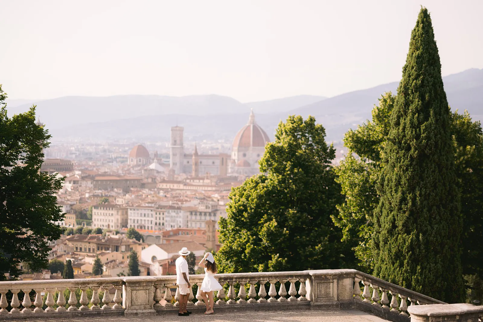 Wide view of Nere and Leo beside a stone balustrade, with Florence Cathedral, rooftops and a tall cypress tree behind them.
