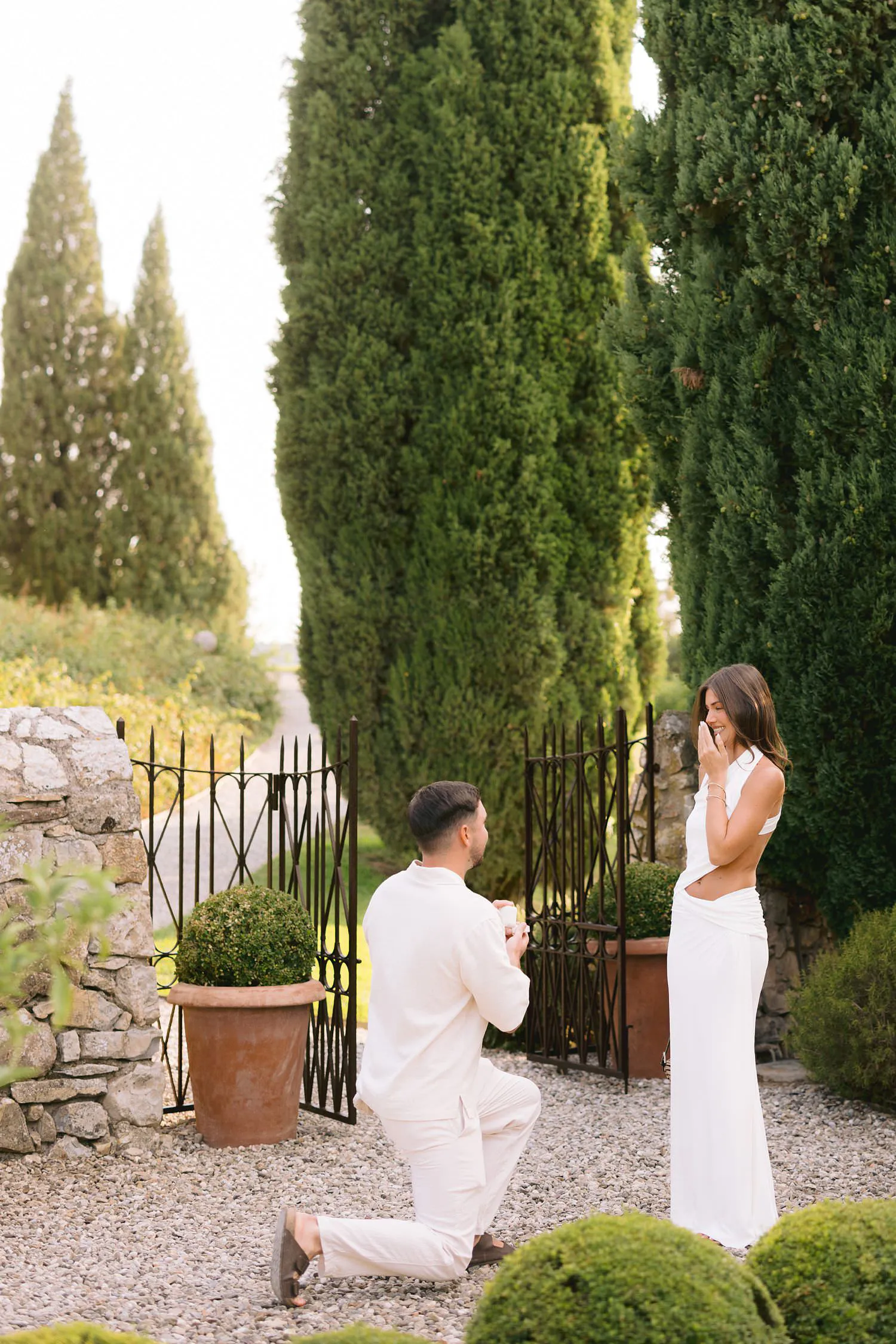 A man kneeling during the surprise proposal as his partner raises her hand to her face in the castle garden.