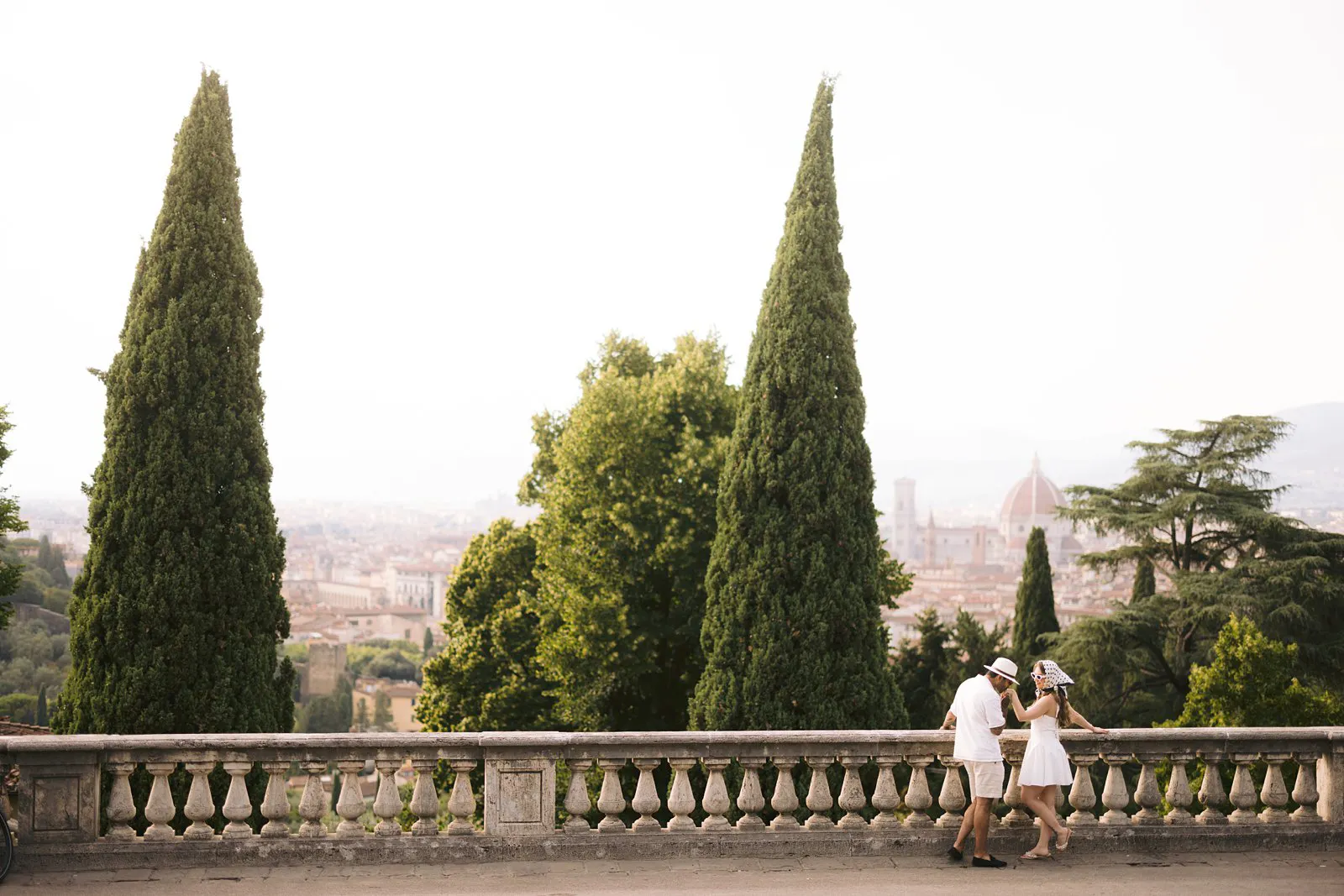 Wide view of Nere and Leo beside a stone terrace balustrade, framed by tall cypress trees above Florence.