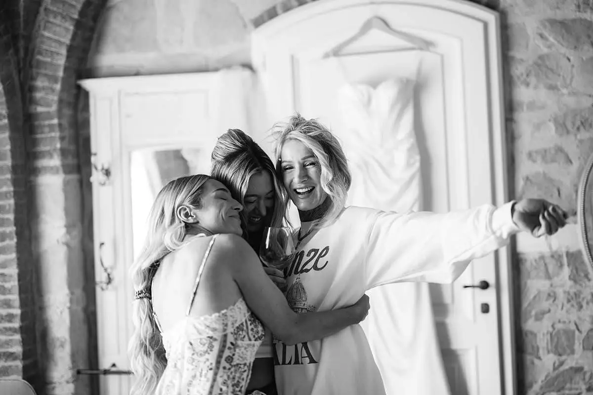 Bride laughs with mother and sister during morning preparations, with her wedding dress hanging behind them.