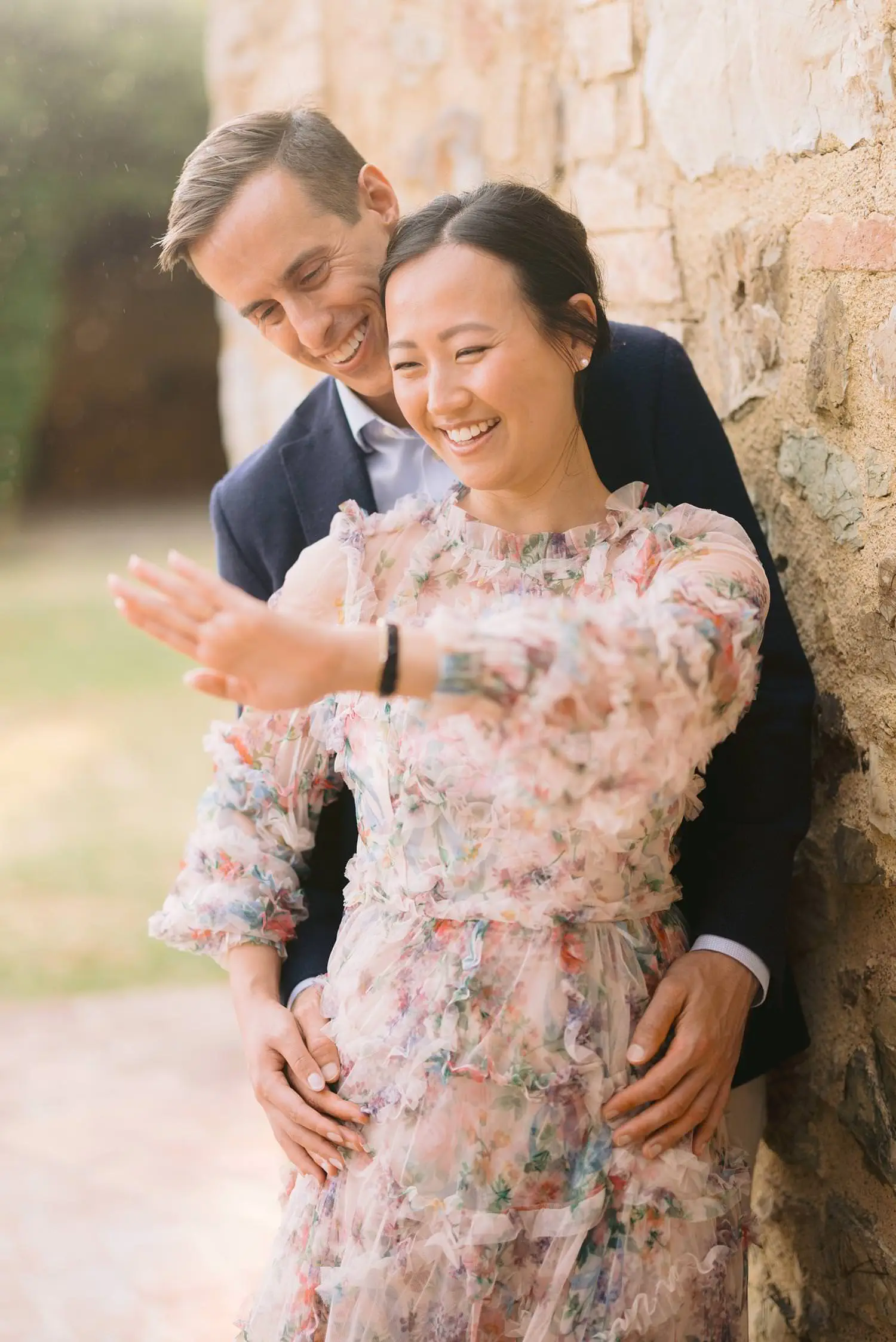 A woman smiles at her engagement ring as her partner embraces her beside the warm stone walls of Capanna House.