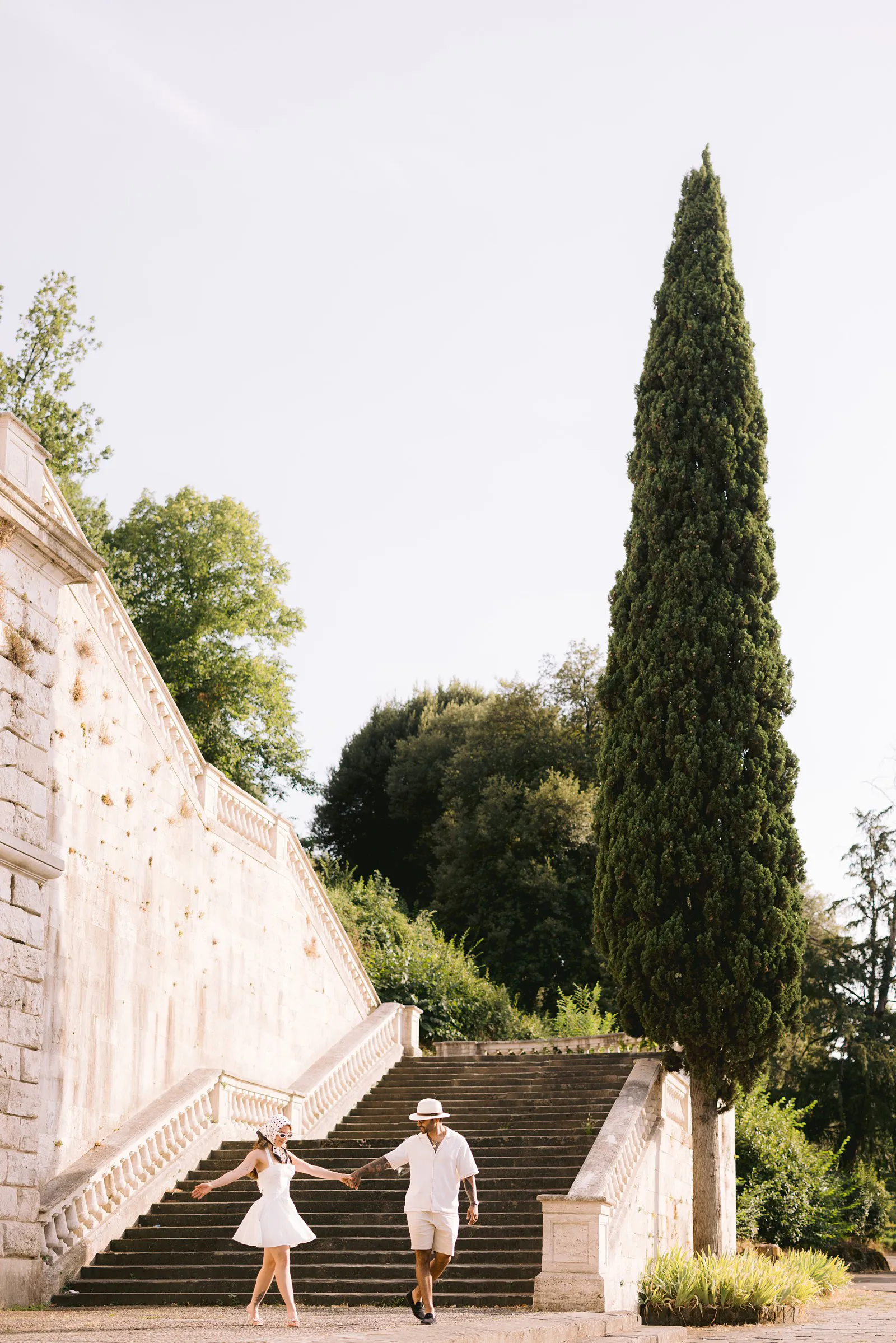 Nere and Leo hold hands as she turns at the foot of a broad stone staircase beside a tall cypress tree in Florence.