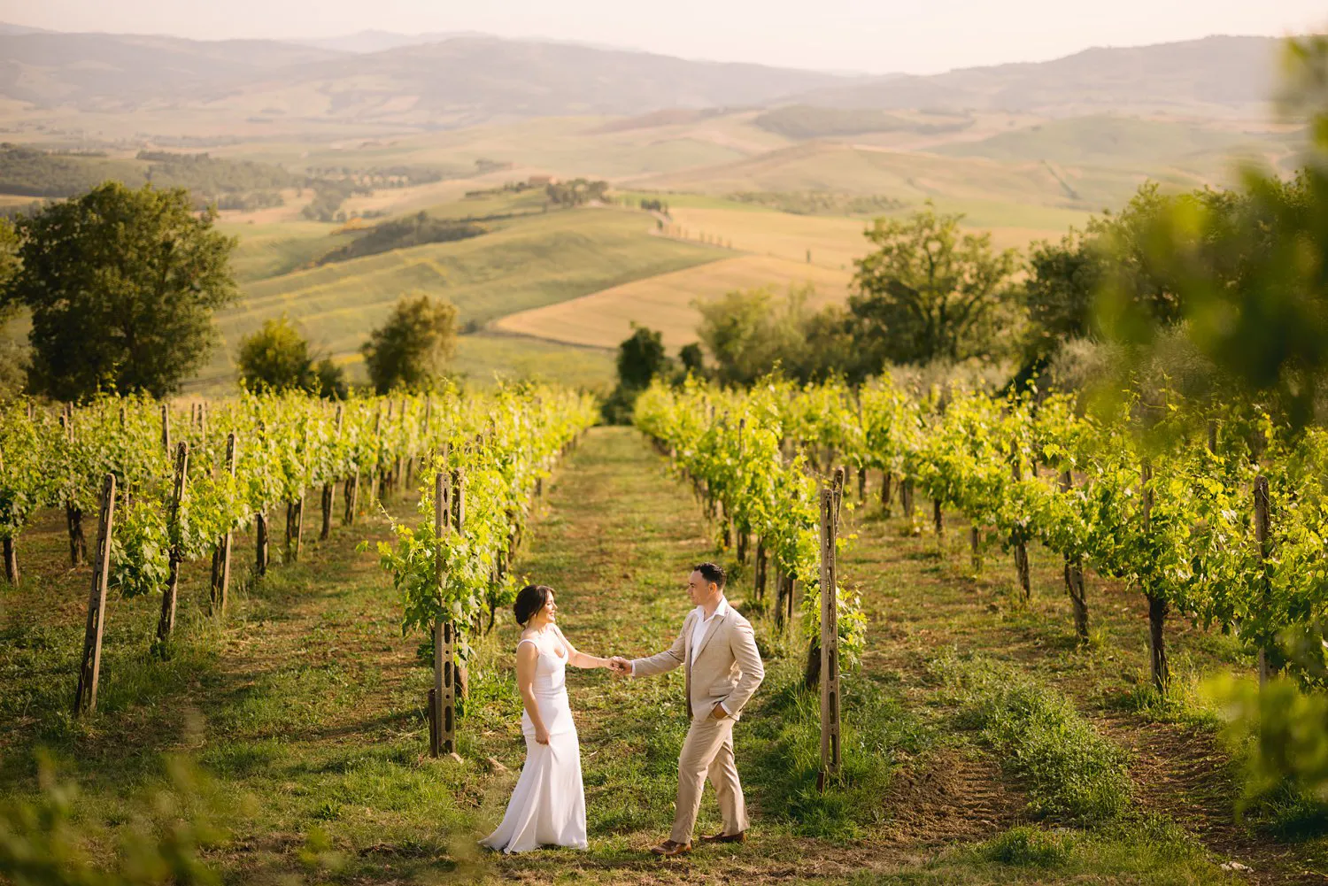 Jessica and Chris hold hands between vineyard rows near Pienza, with the rolling hills of Val d'Orcia behind them.