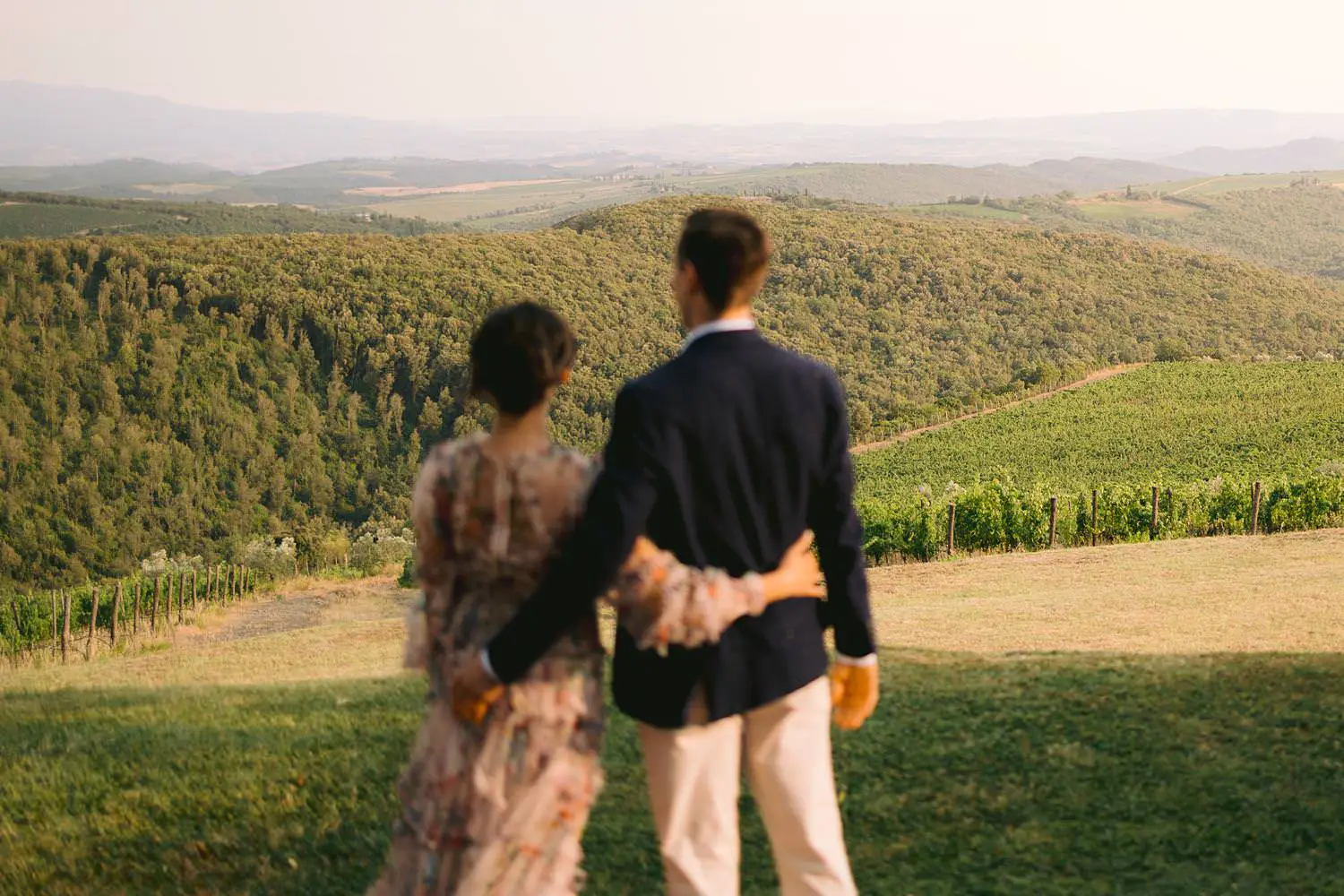 Two partners stand arm in arm, seen from behind against the vineyards and wooded hills of Val d’Orcia.