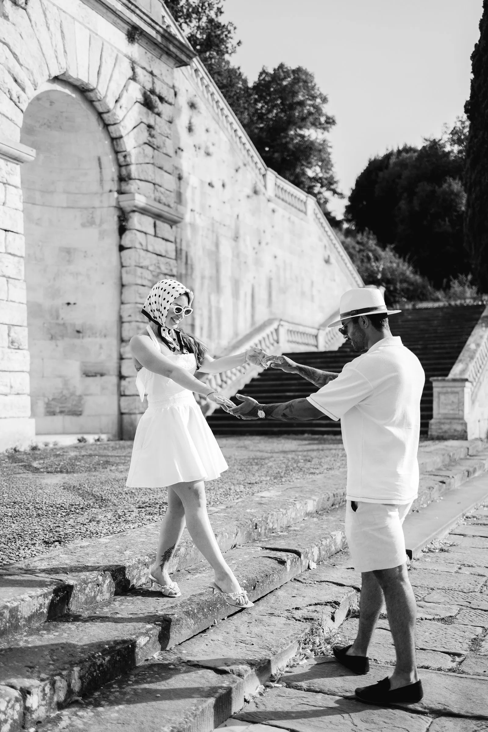 Black-and-white photograph of Leo holding Nere's hands as she steps down beside a stone staircase and arched wall in Florence.