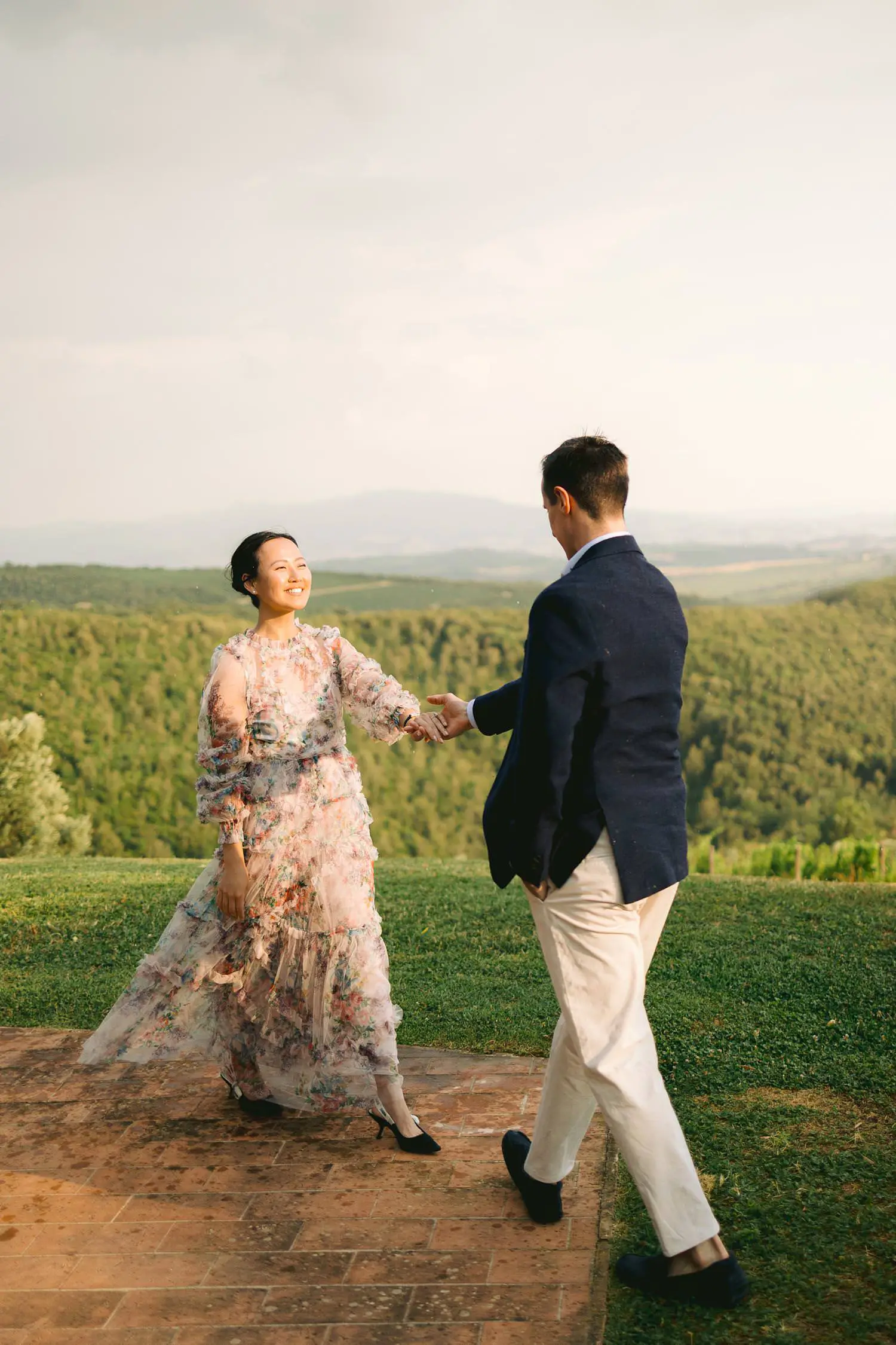 A newly engaged couple turns toward each other, holding hands on the lawn above Capanna’s rolling vineyards.