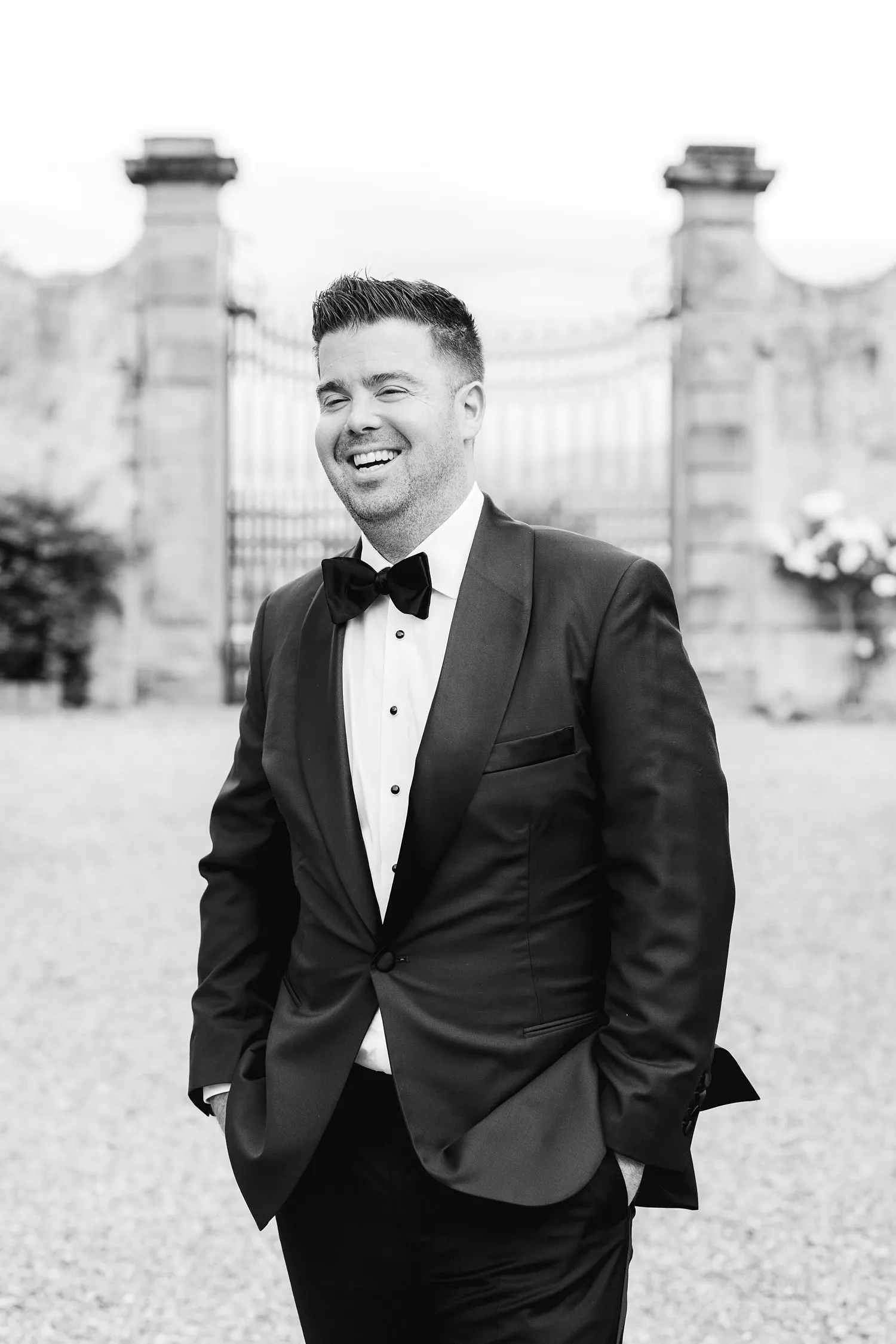 Black-and-white portrait of Kevin smiling in a tuxedo with his hands in his pockets in the gardens of Villa Corsini.