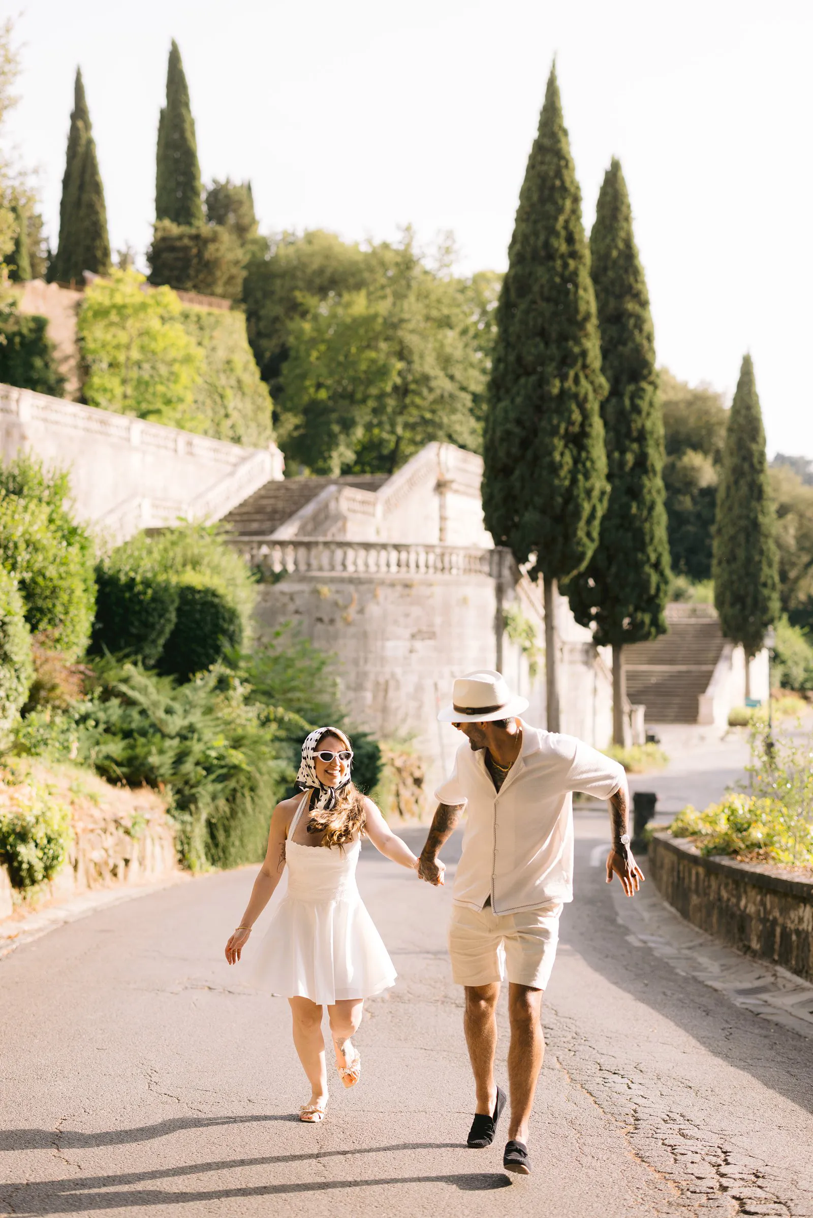 Nere and Leo walk hand in hand along a sunlit road bordered by stone walls, greenery and tall cypress trees in Florence.