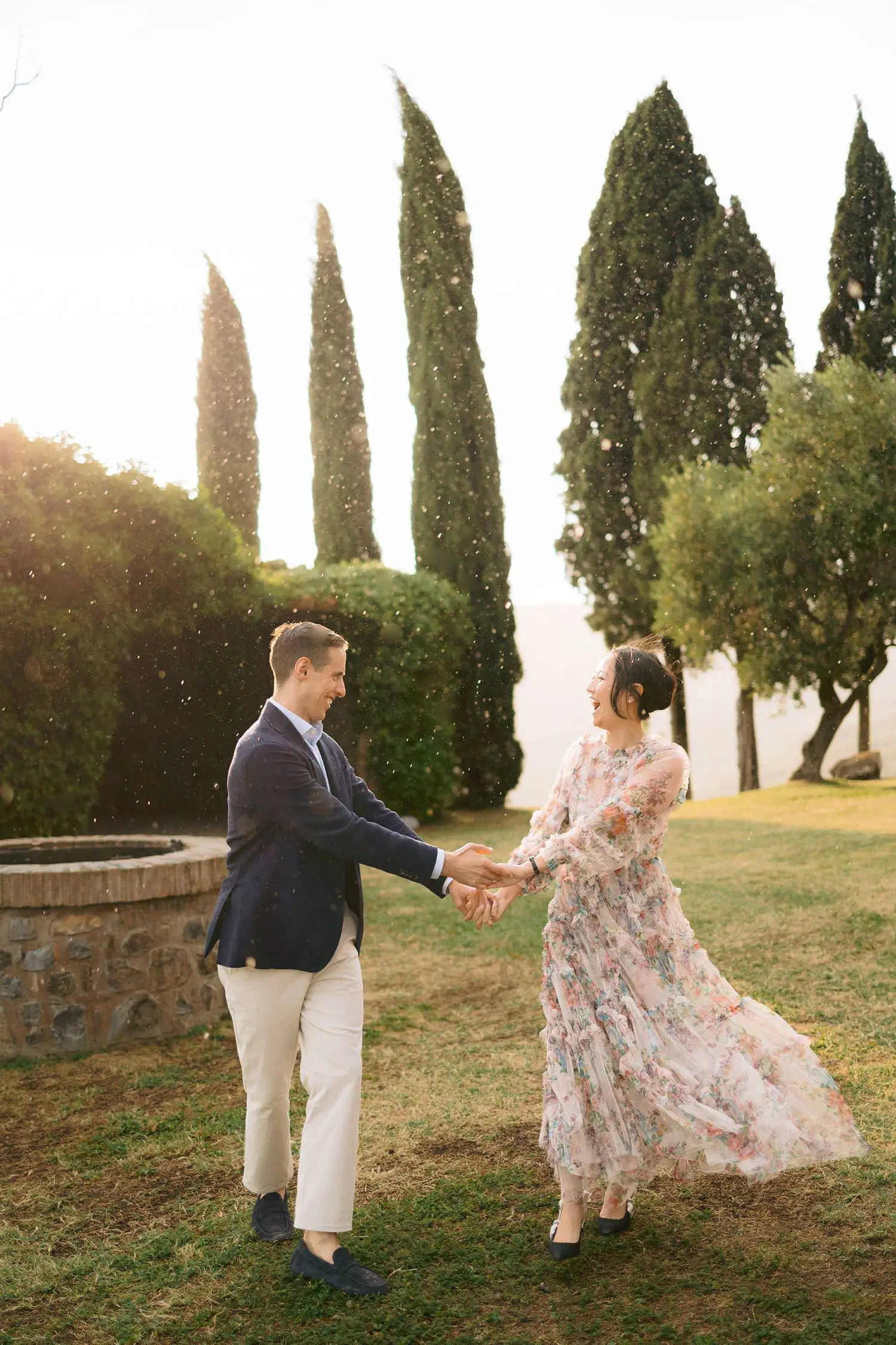 Her floral dress flows as she dances hand in hand with her partner beside Capanna House’s stone well and tall cypresses.