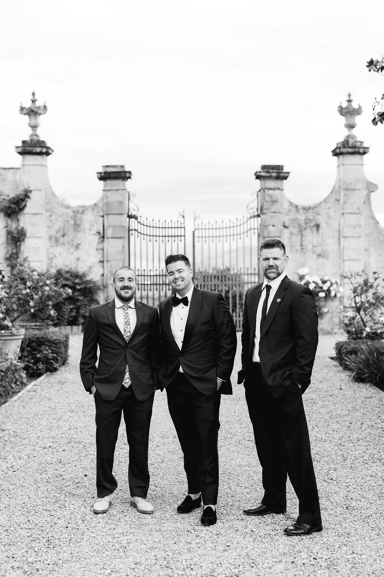 Black-and-white photograph of Kevin standing between two men in dark suits beside the stone garden gates at Villa Corsini.