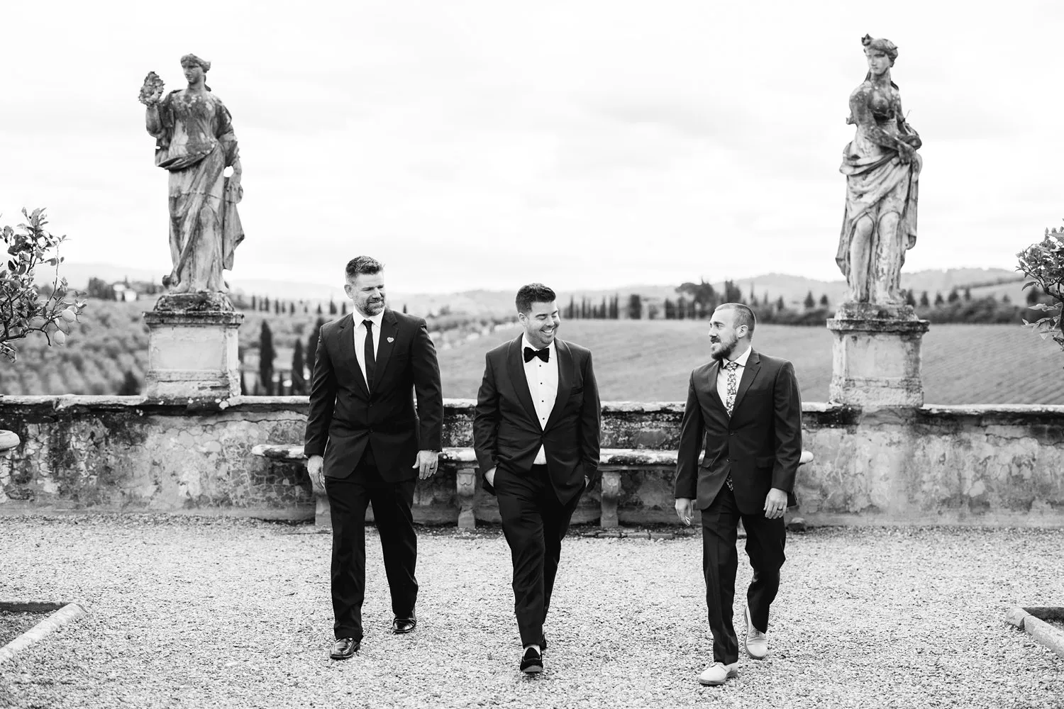 Kevin and two men in dark suits walk across a gravel terrace between stone statues at Villa Corsini, photographed in black and white.
