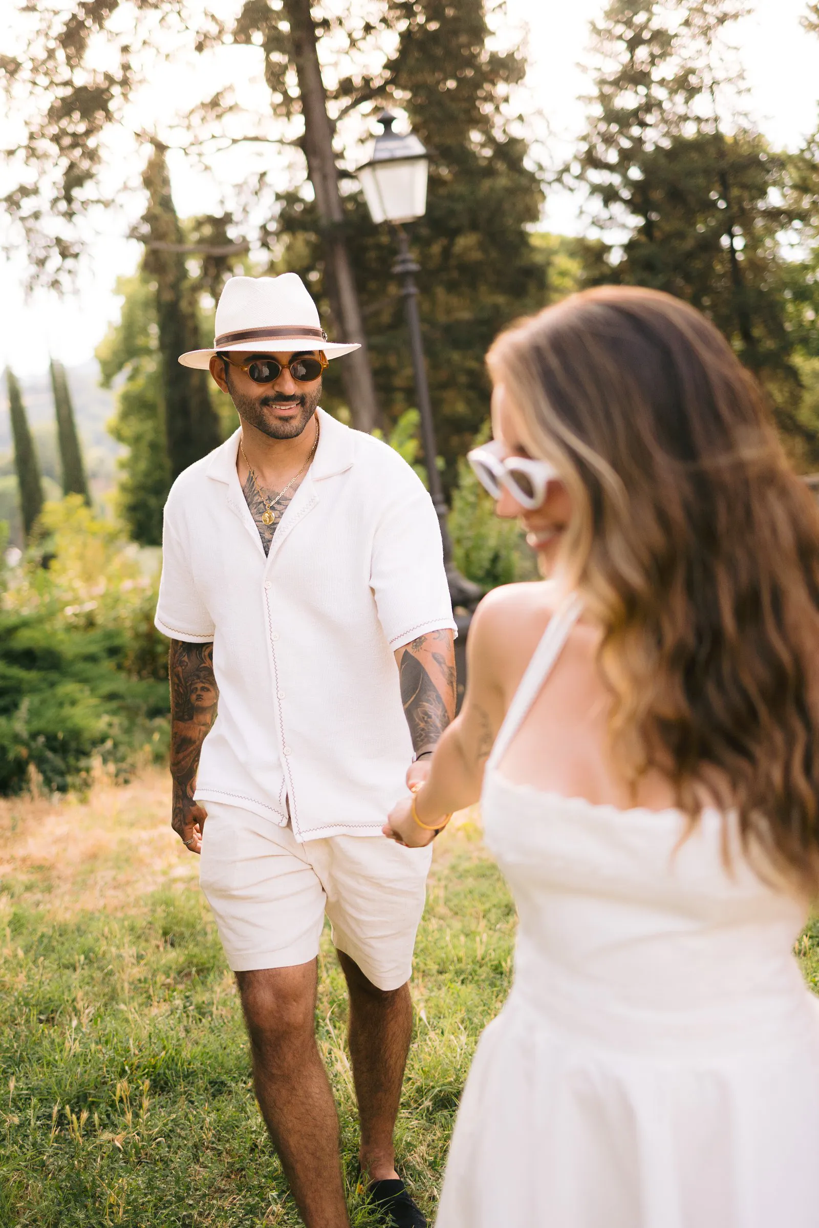 Nere smiles in the foreground while holding Leo's hand, with a garden path and cypress trees behind them in Tuscany.