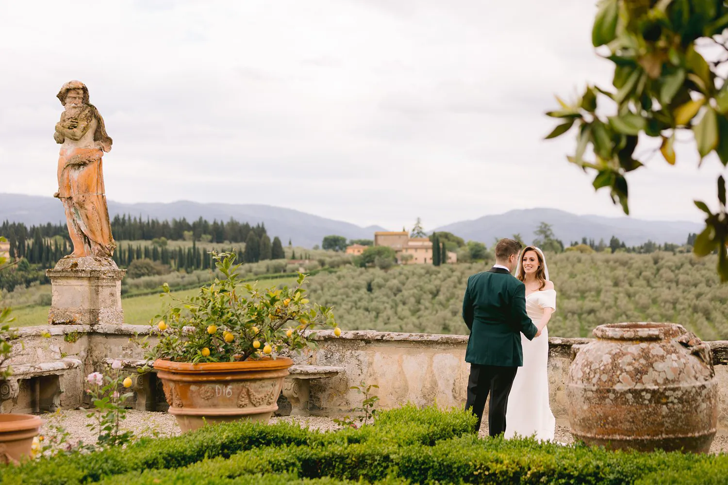 Kathryn and Kevin face each other in a formal garden at Villa Corsini, beside a statue and terracotta pots overlooking the Tuscan hills.
