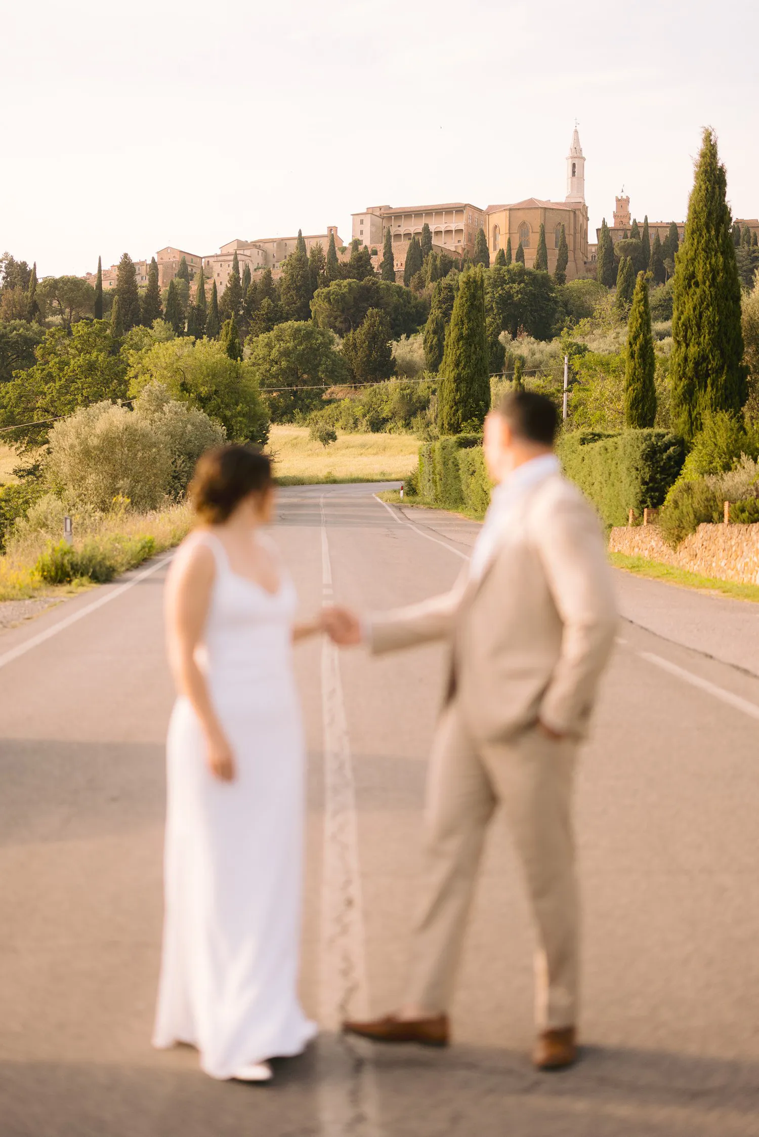 Jessica and Chris hold hands on a country road below Pienza, their figures softly out of focus against the hilltop town.