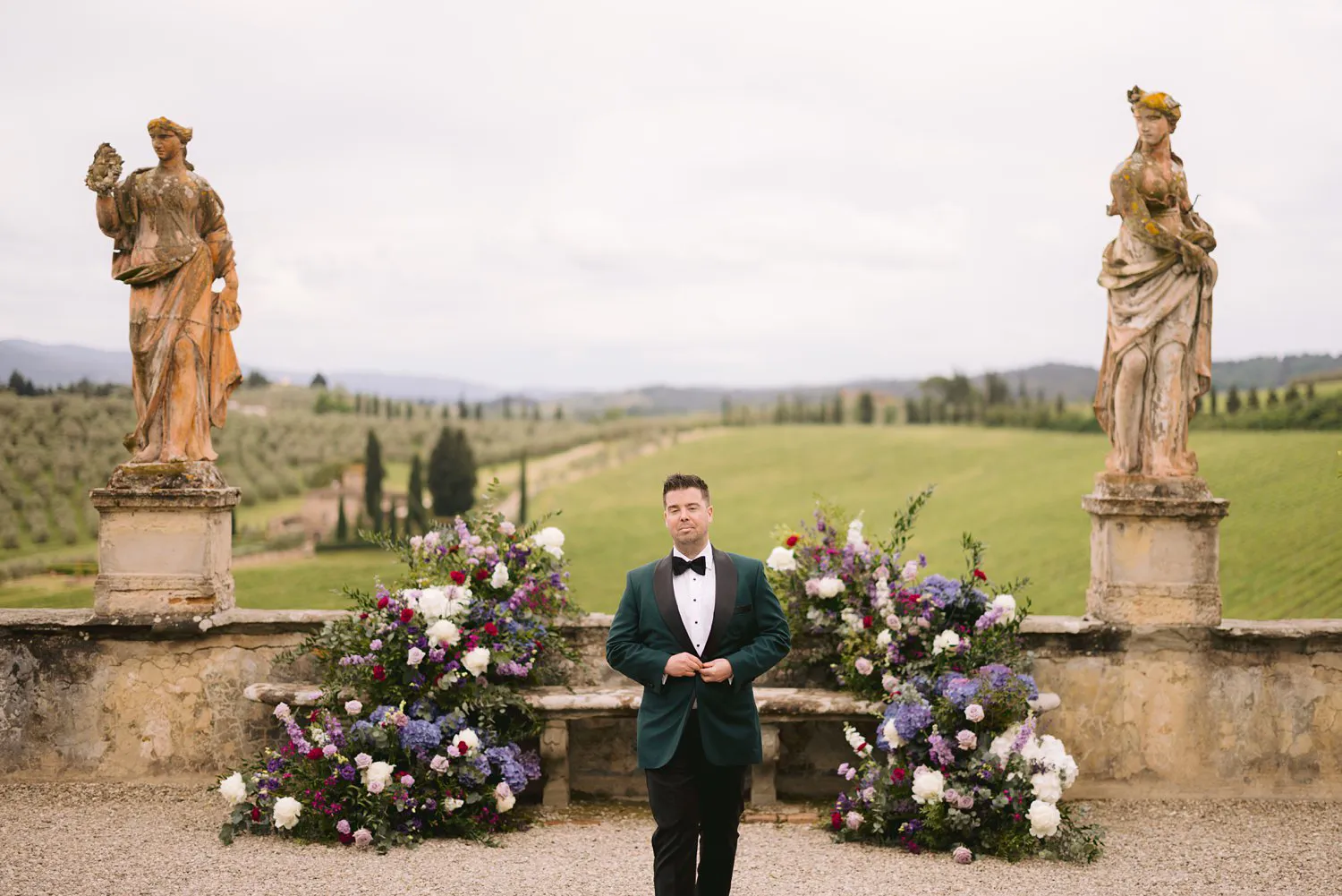 Kevin in a deep green tuxedo stands between purple and white floral arrangements beneath two statues in the ceremony garden at Villa Corsini.