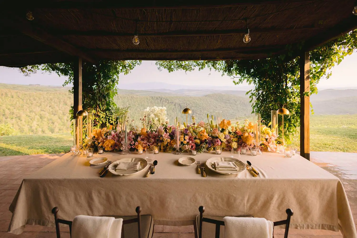 An elegant dinner table filled with pastel flowers beneath Capanna House’s shaded portico, opening onto vineyards and Tuscan hills.
