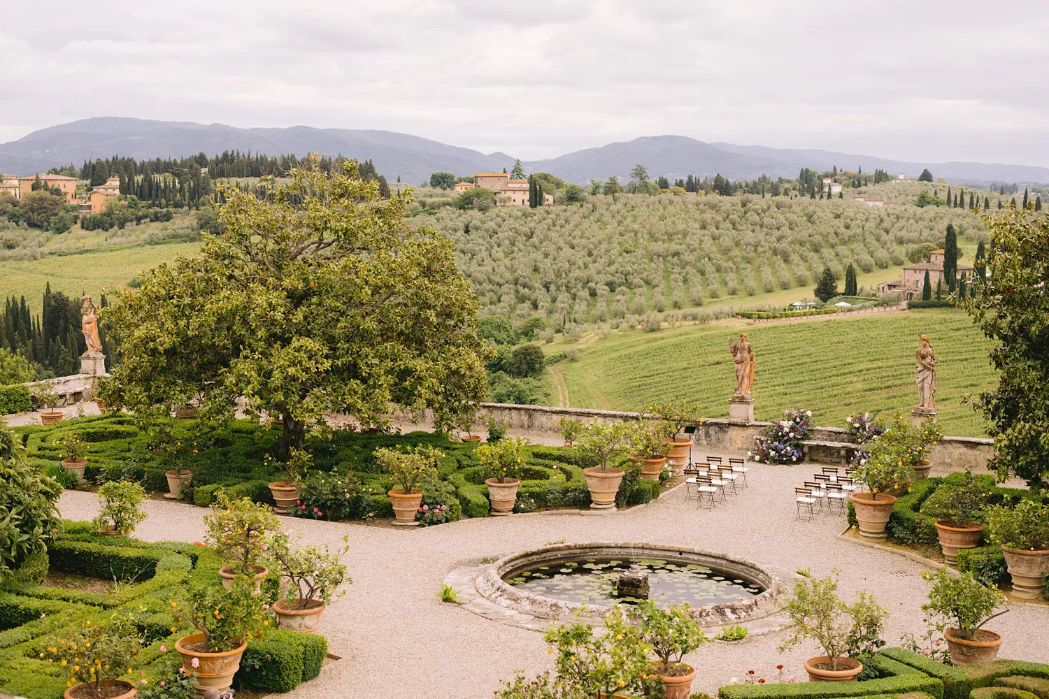 Elevated view of Villa Corsini's formal garden, circular fountain and ceremony chairs, with olive groves and rolling Tuscan hills beyond.