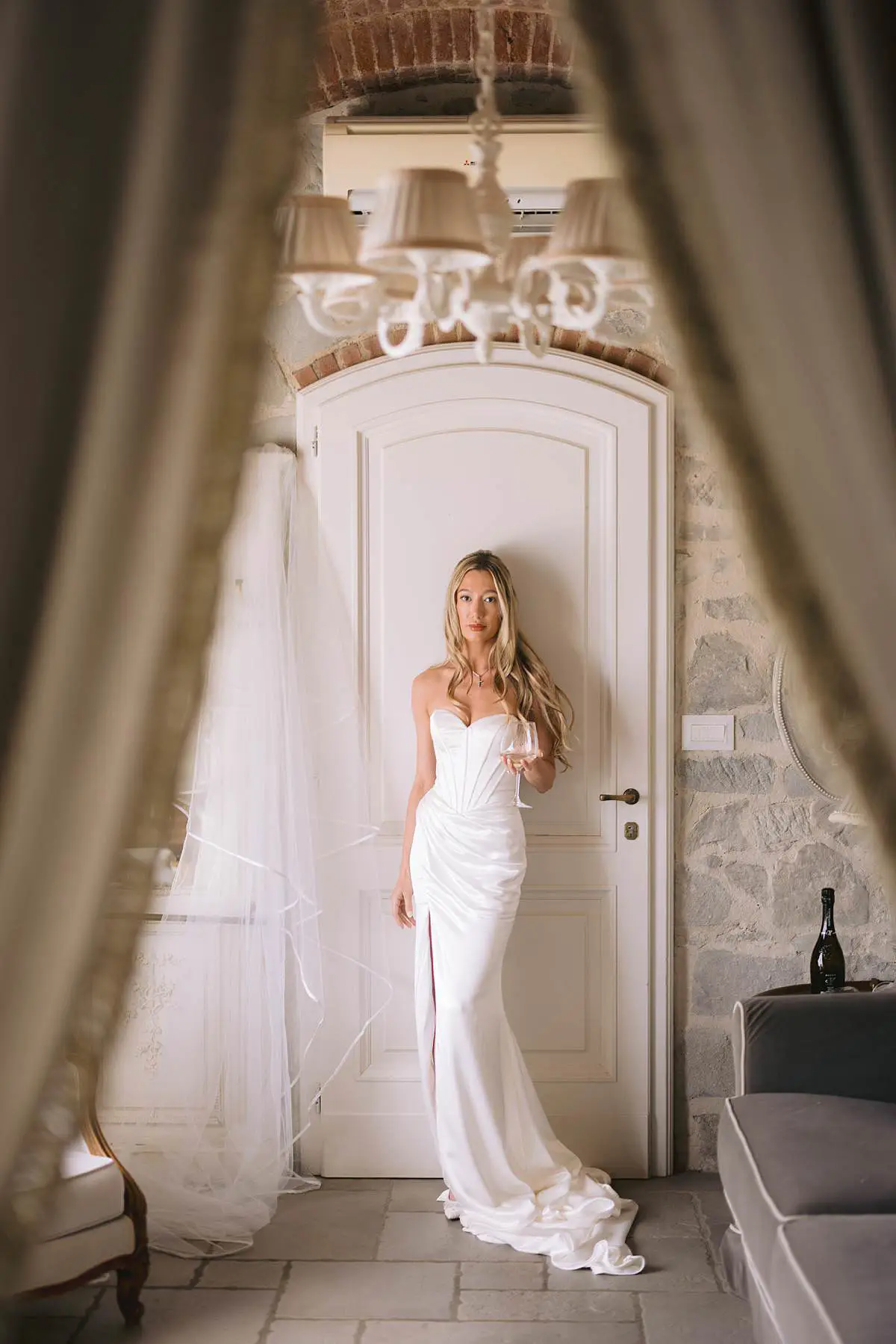 Bride in a white wedding dress holds a glass beside her veil in a Villa Le Fontanelle suite.