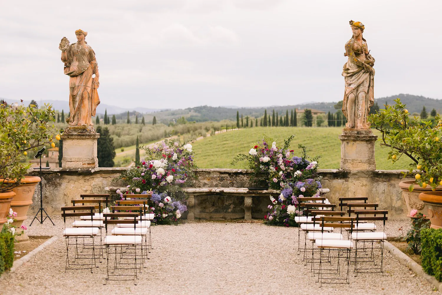 Rows of wooden ceremony chairs face purple and white floral arrangements between stone statues in Villa Corsini's garden overlooking the evocative rolling hills.