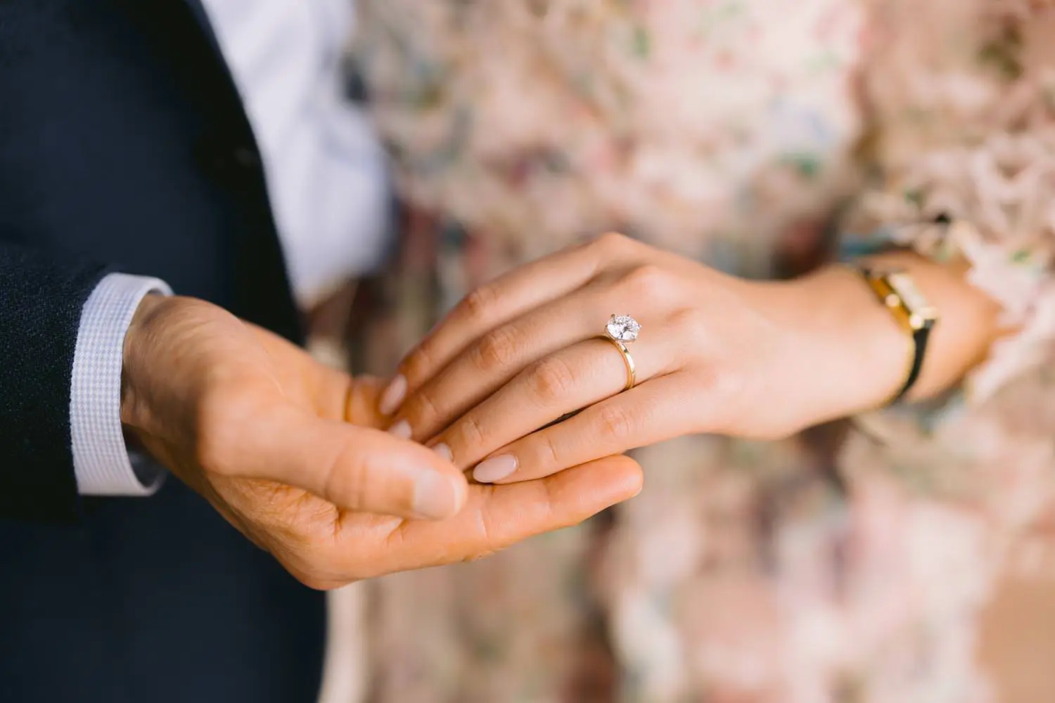 A close-up of joined hands reveals the engagement ring against the soft colours of a floral dress.