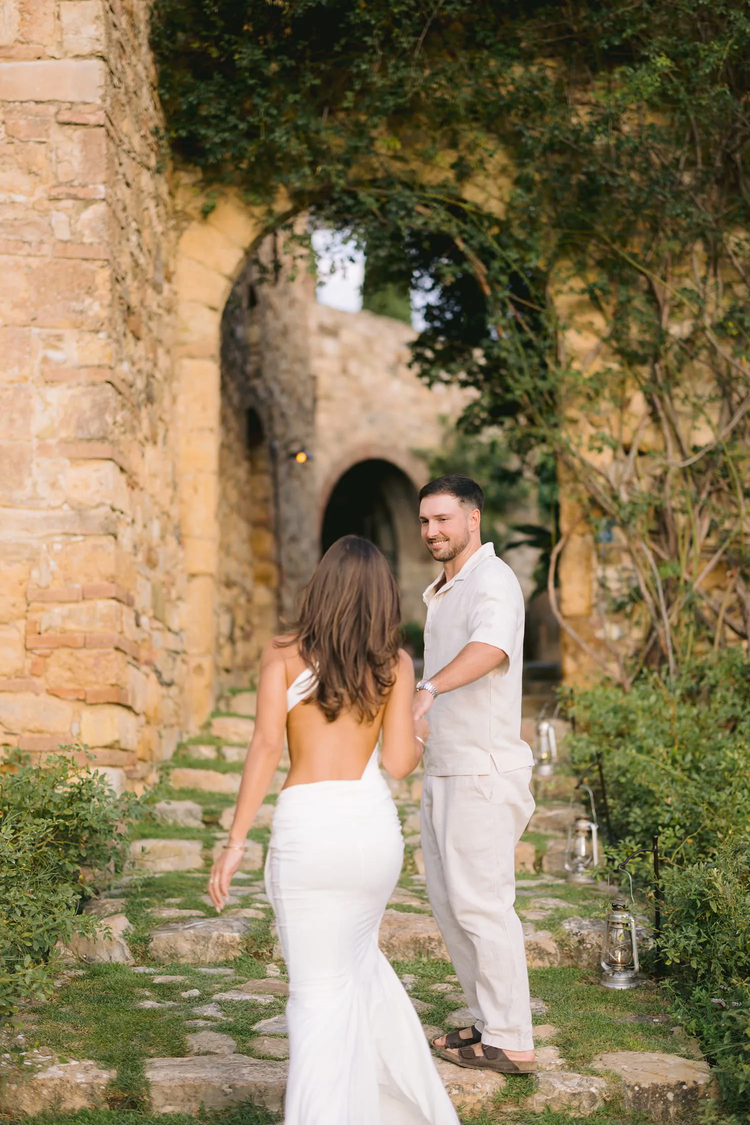 The couple holding hands as they walk up a stone stairway beneath a leafy arch.