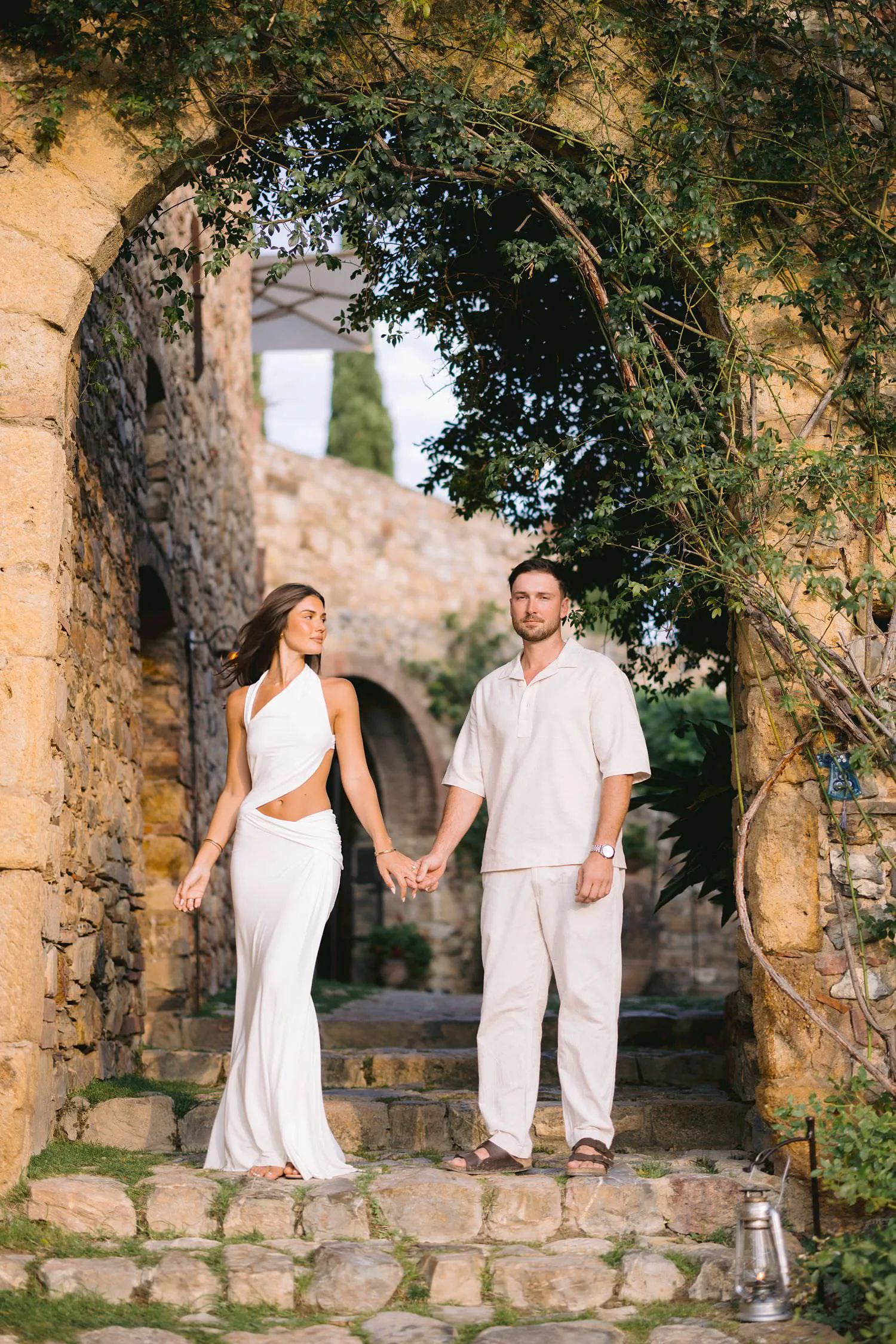 Dekota and Aiden standing hand in hand beneath a stone arch covered with greenery.
