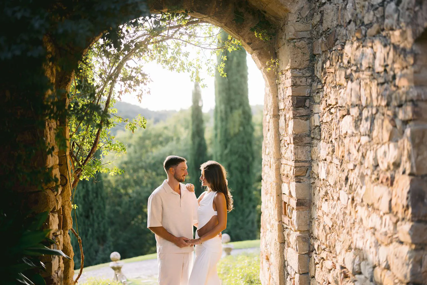 Dekota and Aiden facing each other beneath a shaded stone archway in warm light.