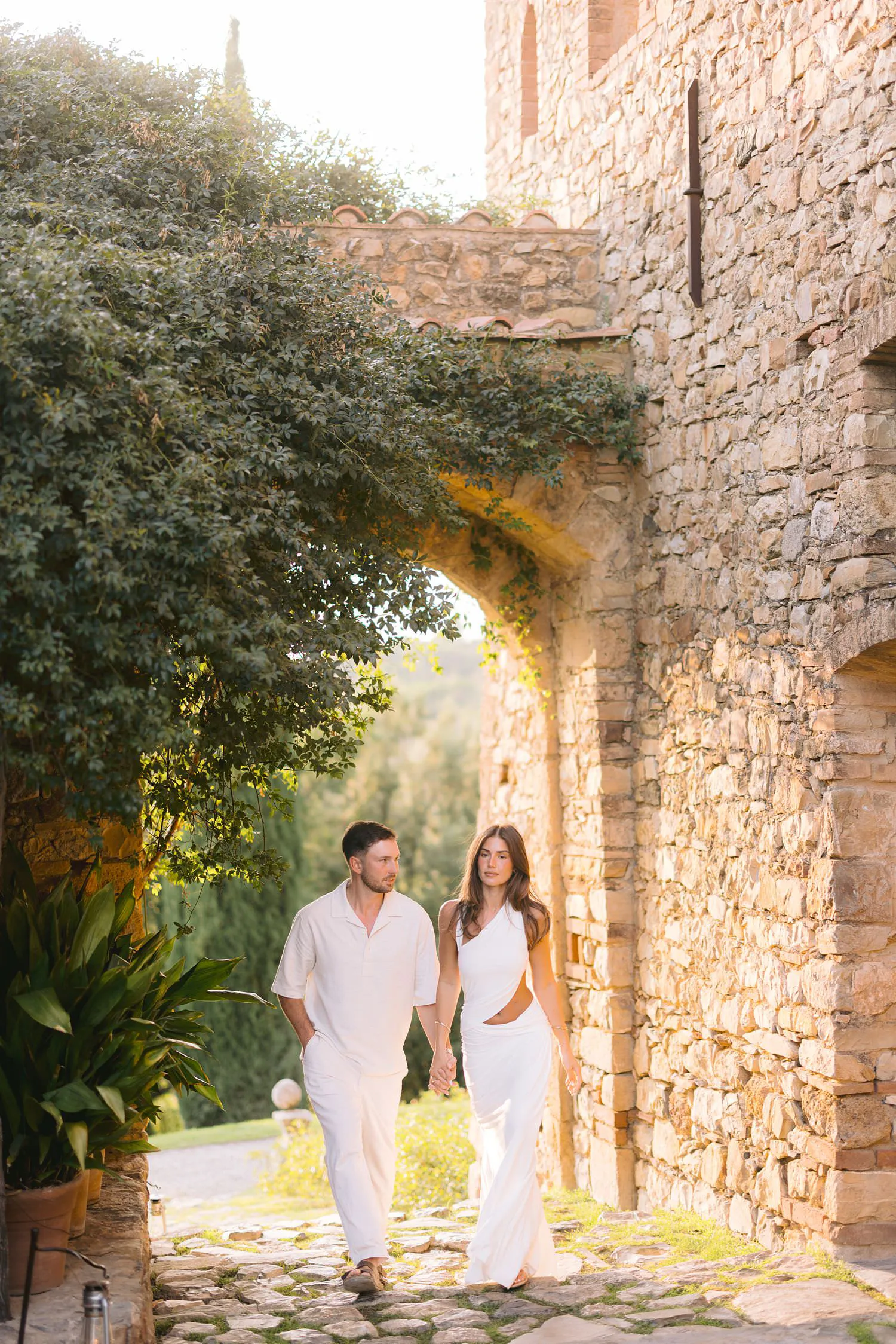Dekota and Aiden walking hand in hand through a stone courtyard beneath an arch.