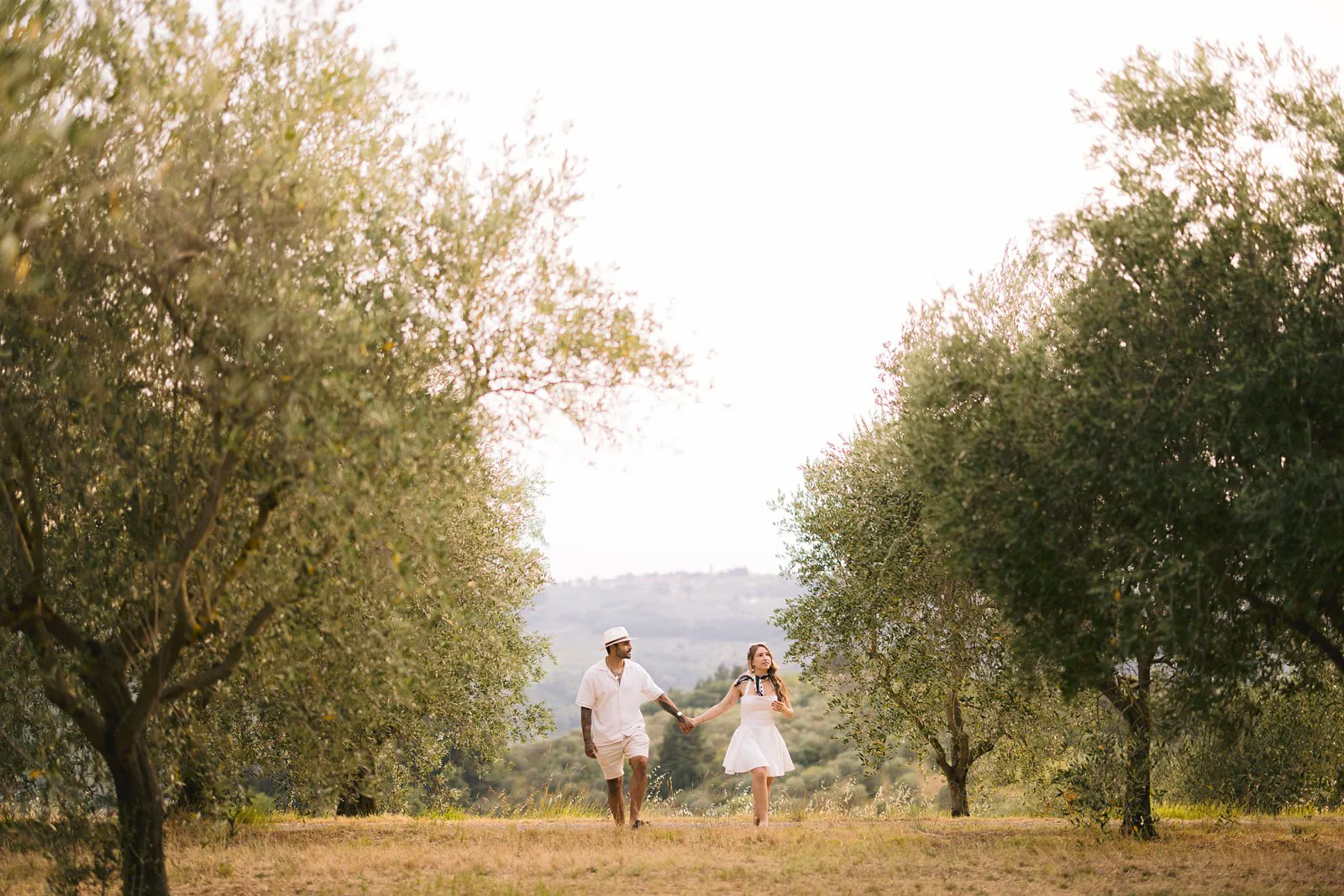 Wide photograph of Nere and Leo walking hand in hand across dry grass between olive trees in the Chianti countryside.