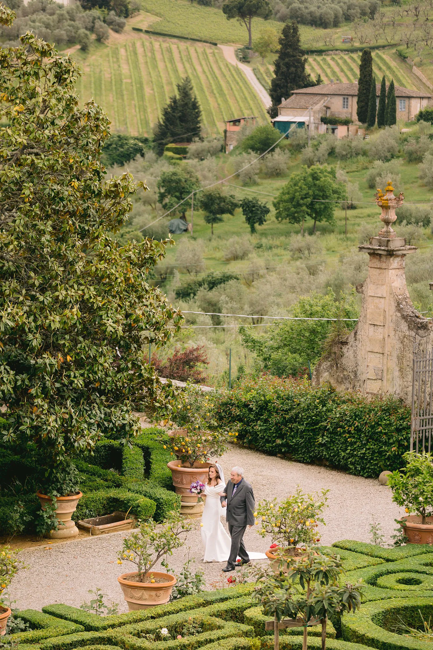 Kathryn walks arm in arm with a man in a grey suit through Villa Corsini's formal garden, beneath a hillside of vineyards and olive trees.
