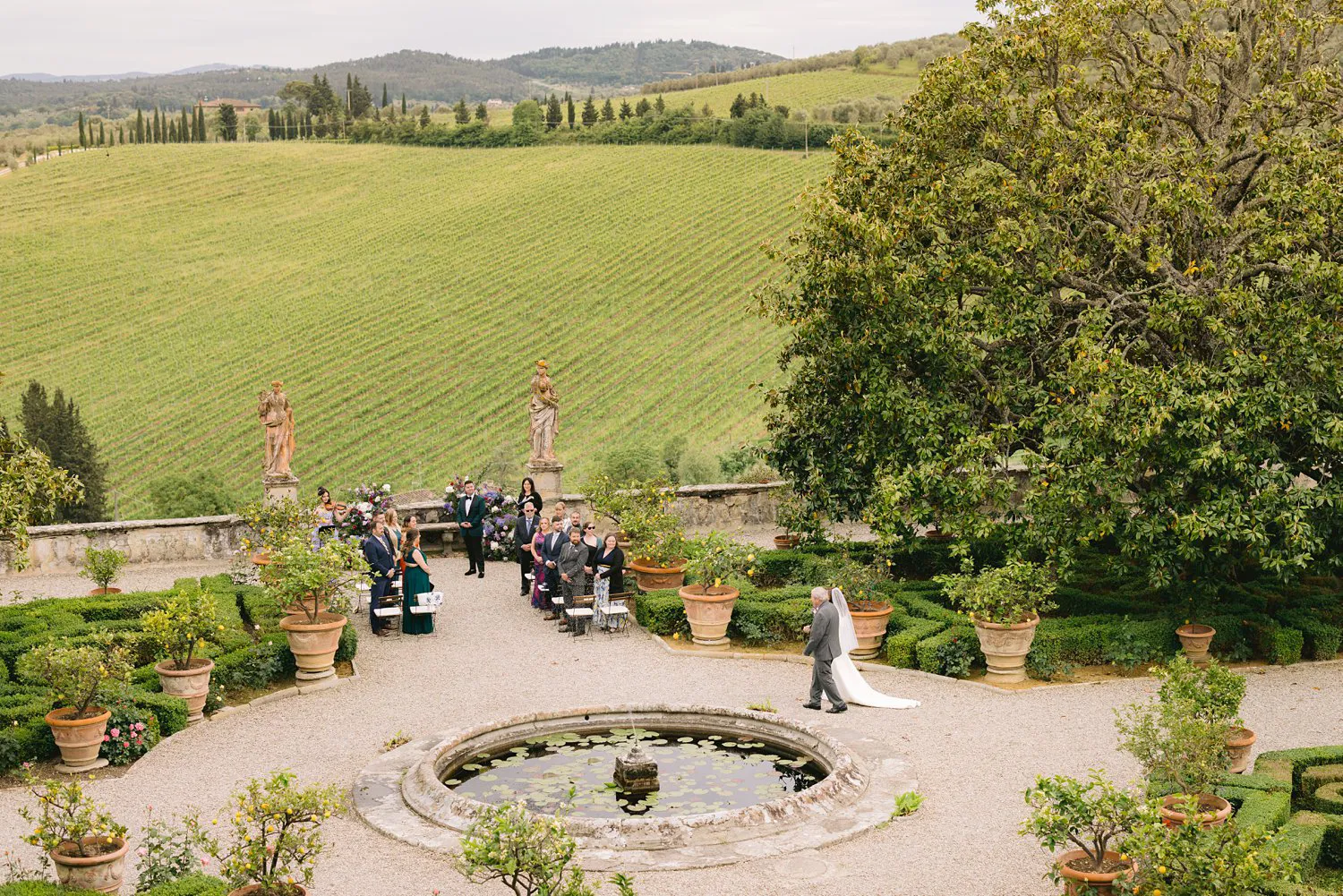 Elevated view of Kathryn entering Villa Corsini's ceremony garden with her escort, as guests stand and Kevin waits beyond a circular lily pond.