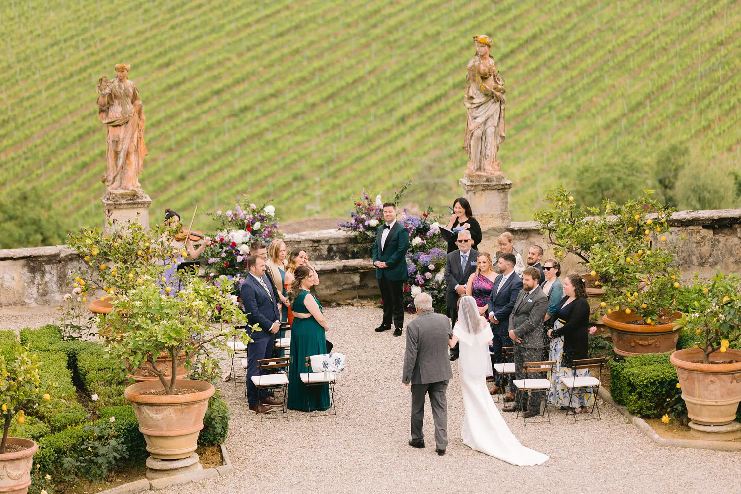 Guests stand beside the aisle as Kathryn and her escort approach Kevin at Villa Corsini, with a violinist, flowers and two statues behind the groom.