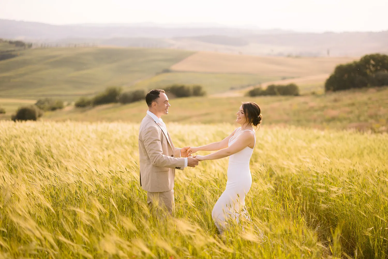 Chris holds Jessica's hands as she leans back among tall sunlit stems, with the hills of Val d'Orcia behind them.