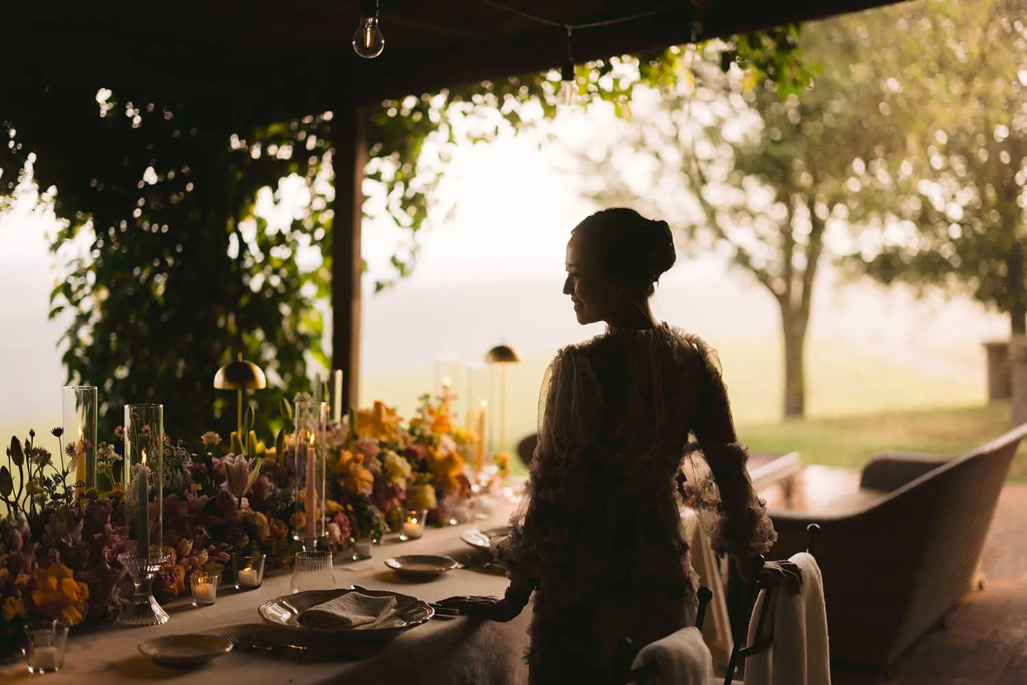 A woman’s silhouette beside the flower-filled dinner table at Capanna House, framed by the shaded portico and luminous Tuscan countryside.