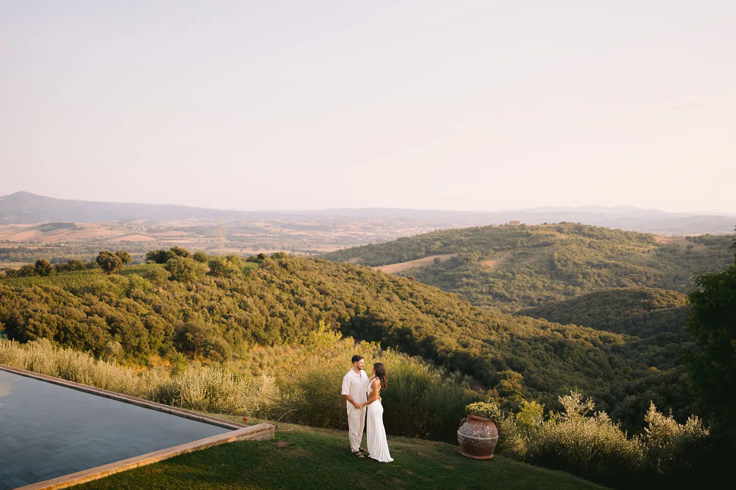 Wide romantic view of Dekota and Aiden beside the pool with rolling Tuscan hills stretching behind them.