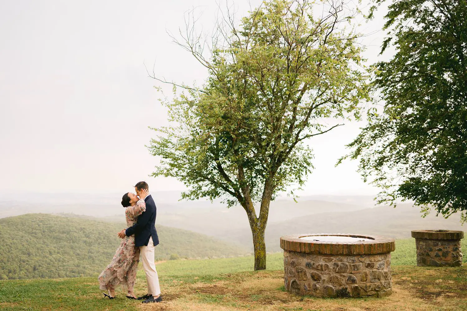 A couple shares an embrace beside a tree and stone well at Capanna House, with the soft Tuscan landscape stretching beyond.