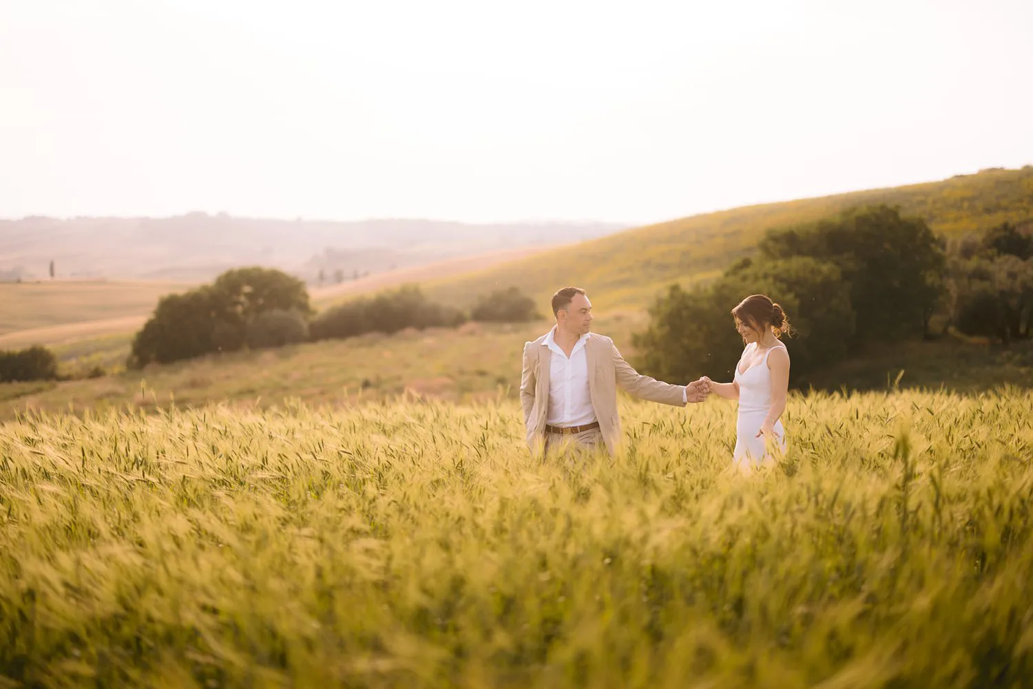 Jessica and Chris walk hand in hand through tall grain near Pienza, with warm light falling across the surrounding hills.