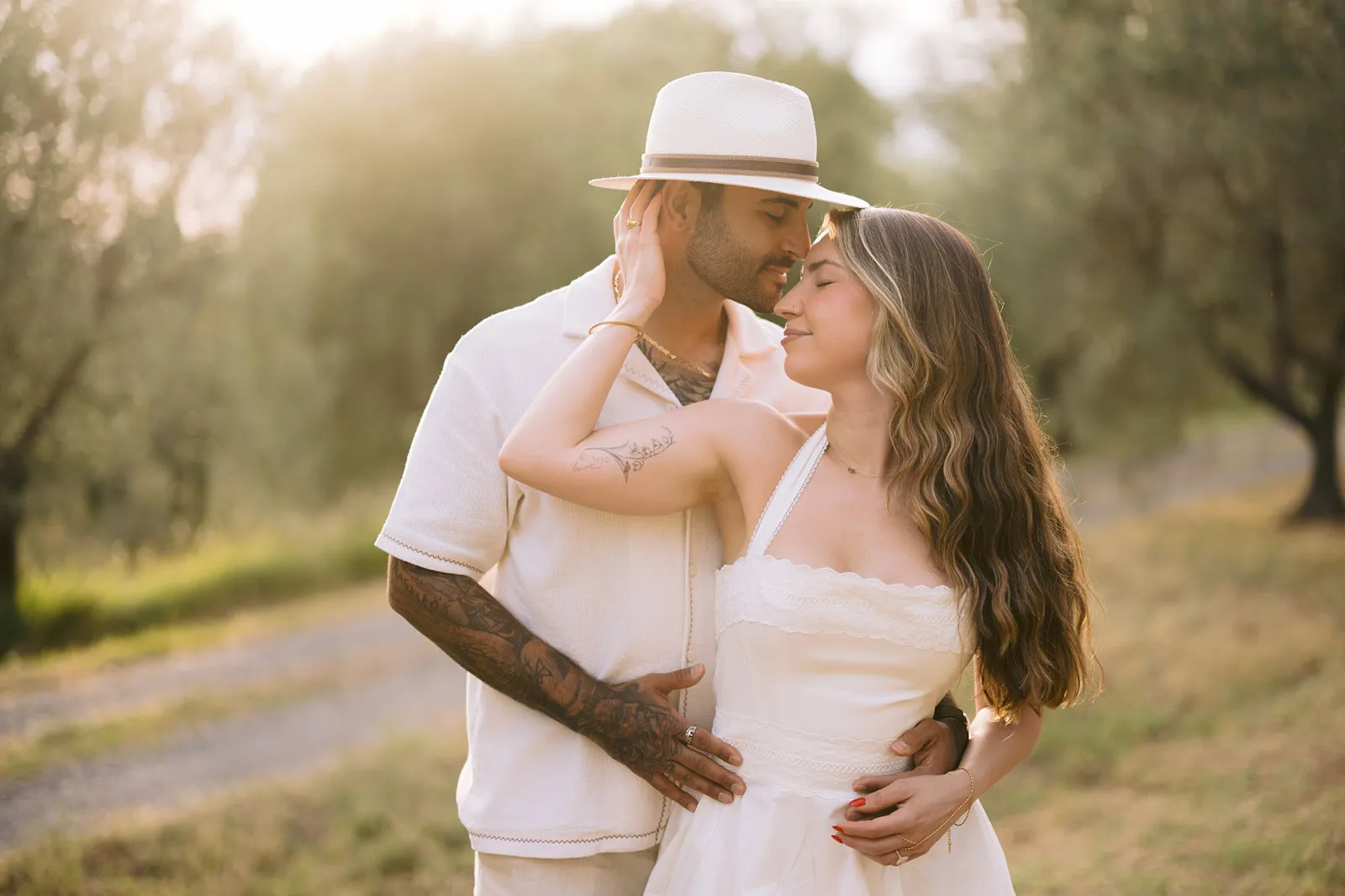 Nere and Leo embrace beneath olive trees in Chianti, with warm backlight and a country path behind them.