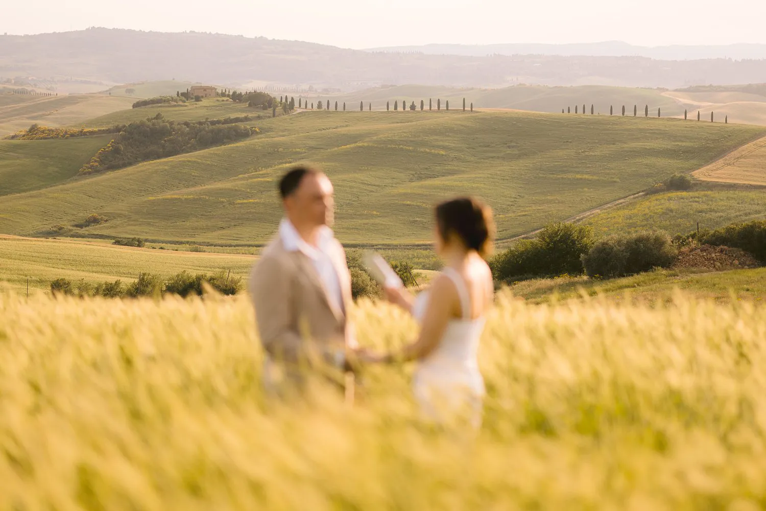 Jessica and Chris stand facing each other during their vow renewal, softly out of focus beyond sunlit grain near Pienza.