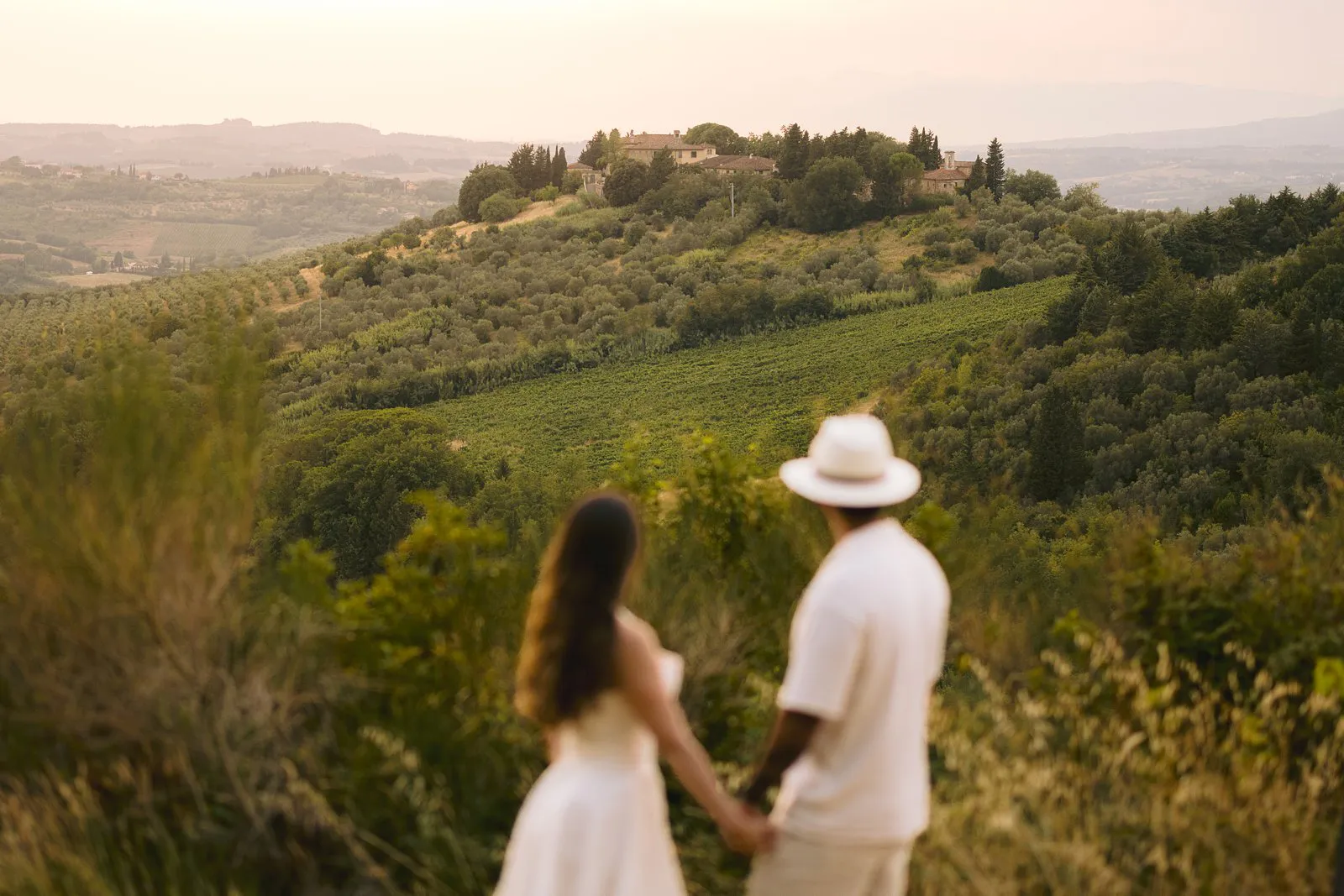 Nere and Leo seen from behind holding hands, softly out of focus before Chianti vineyards and hilltop buildings in evening light.
