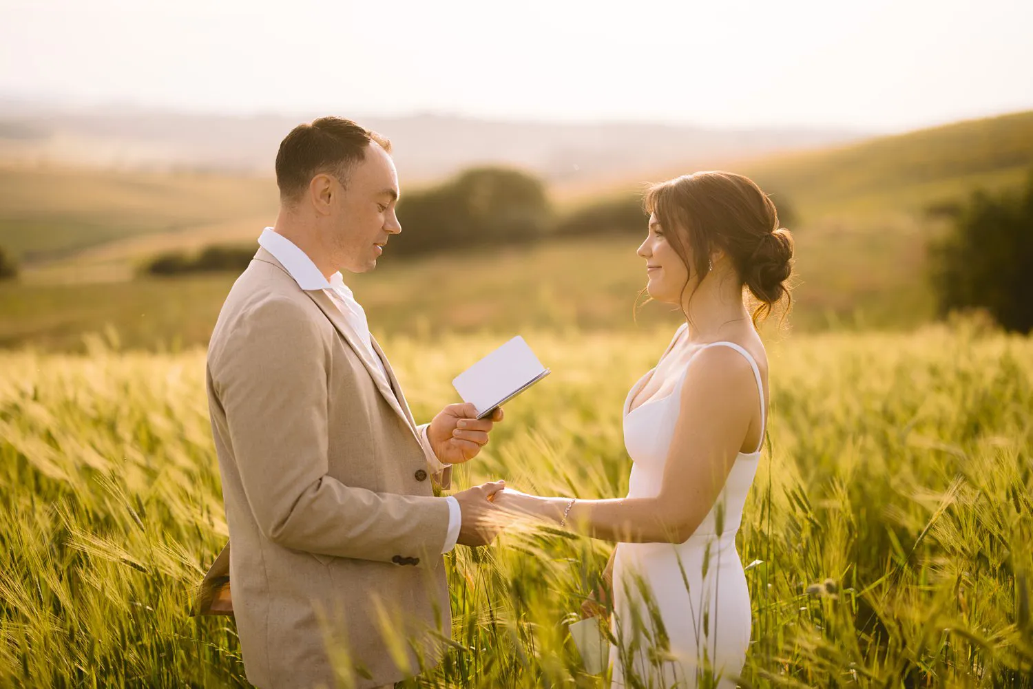 Chris reads his written vows while holding Jessica's hand in a sunlit field near Pienza during their vow renewal.