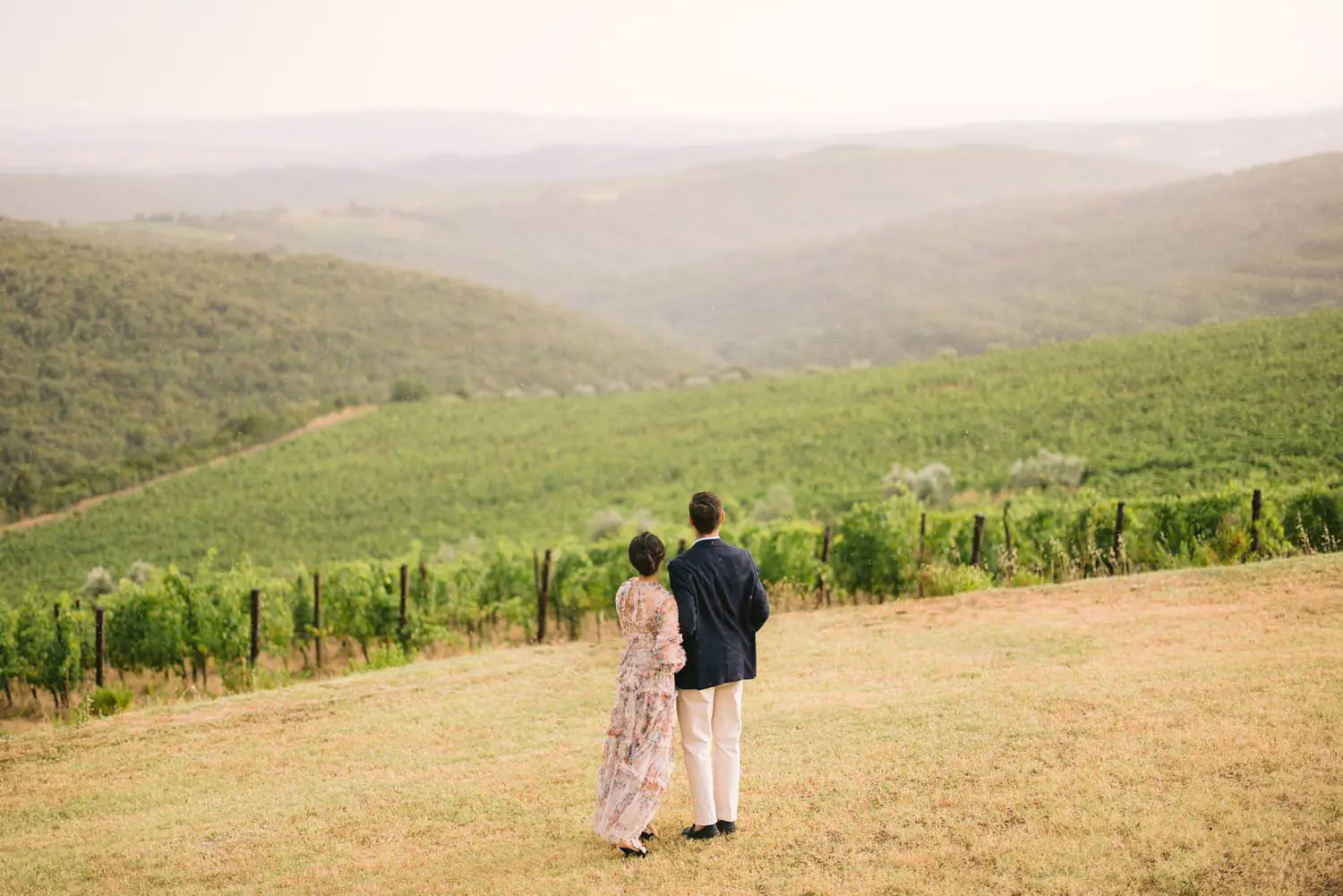 A wide engagement portrait frames two partners overlooking vineyard rows and wooded hills at Castiglion del Bosco in Val d’Orcia.