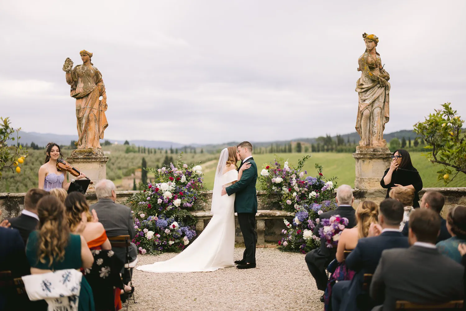 Kathryn and Kevin kiss during their garden ceremony at Villa Corsini as seated guests applaud, with a violinist beside the floral arrangements.