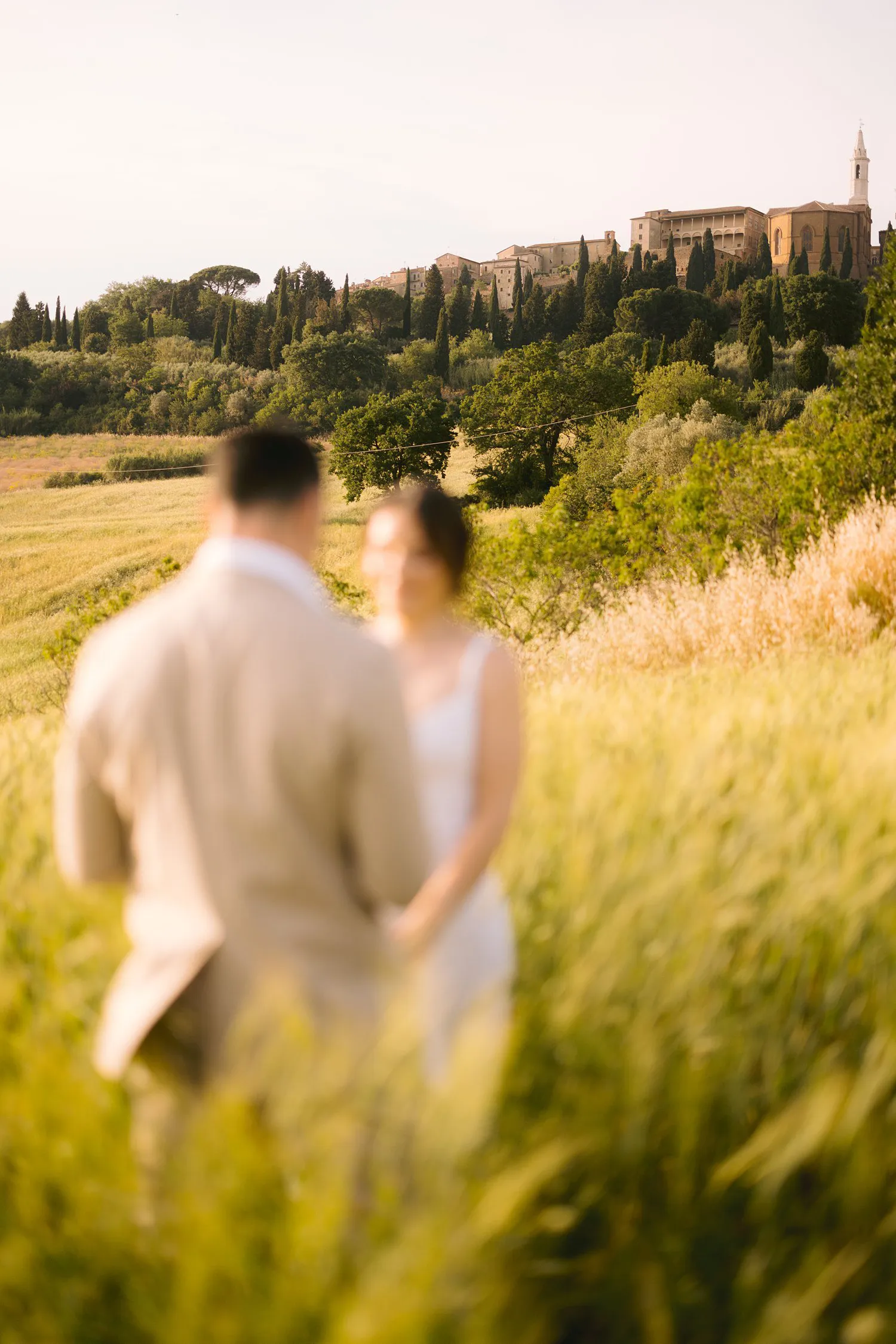 Jessica and Chris appear softly out of focus during their vows, framed by tall grain with Pienza on the hill beyond.