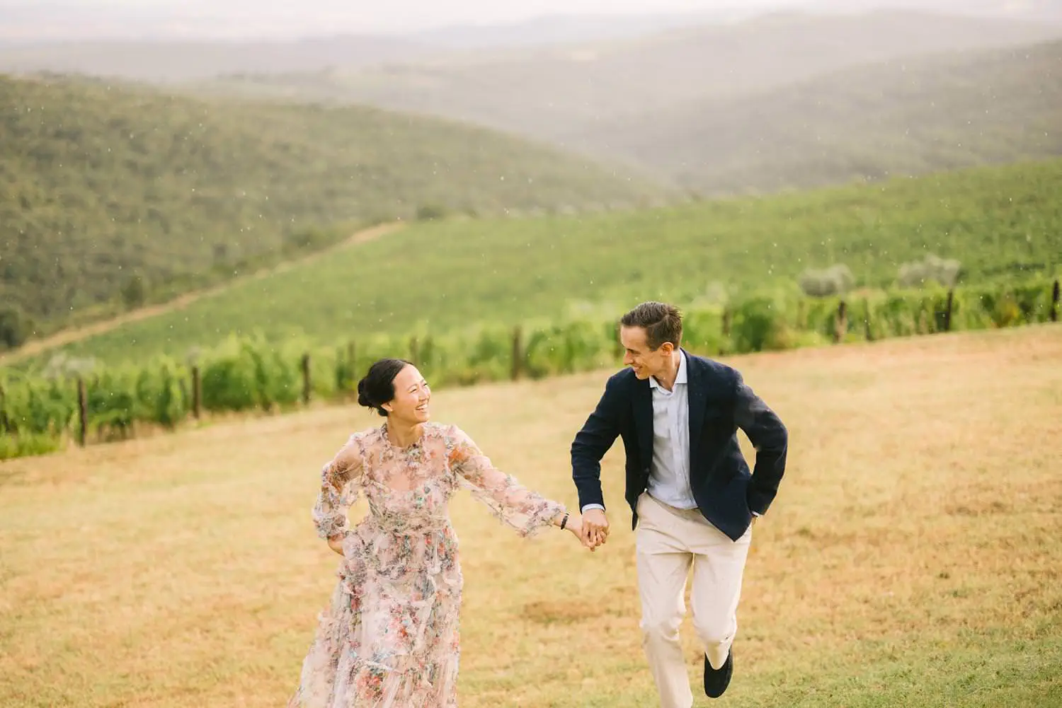 A couple laughs and skips hand in hand across the grass above Capanna’s vineyard rows during their engagement portraits.